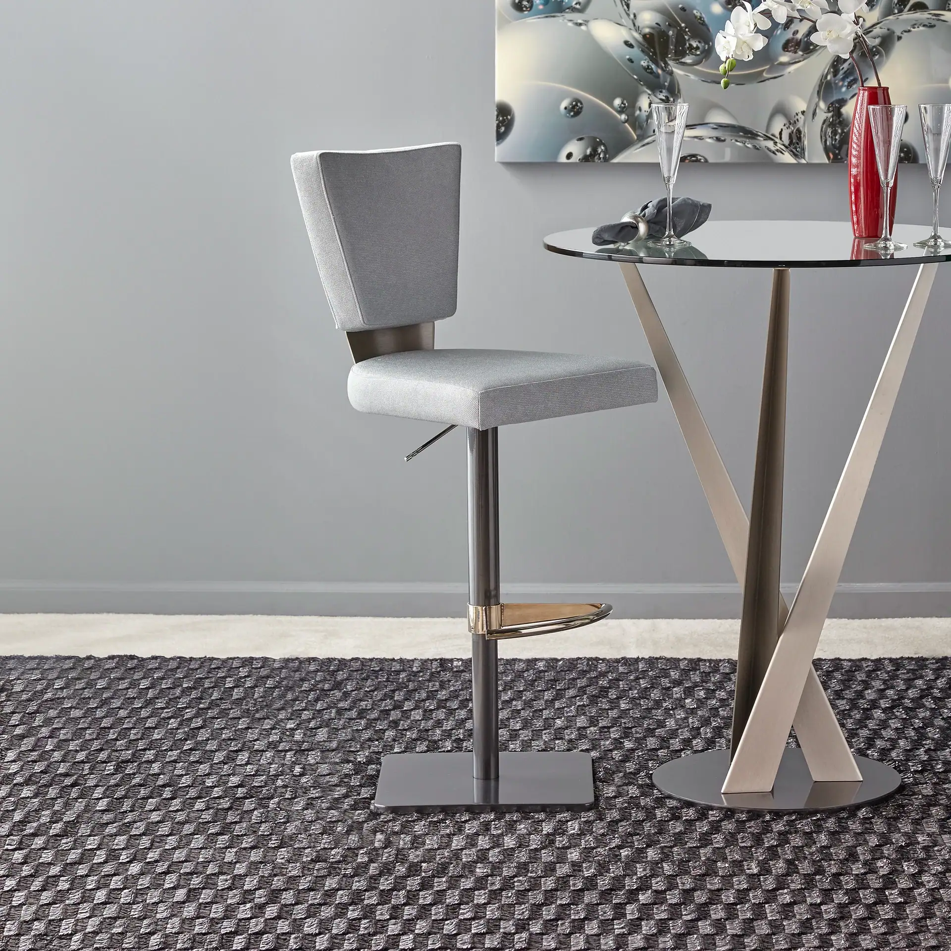 A modern gray upholstered bar stool and a round glass-top bar table with champagne flutes and white orchids, set against a gray wall with abstract metallic art.