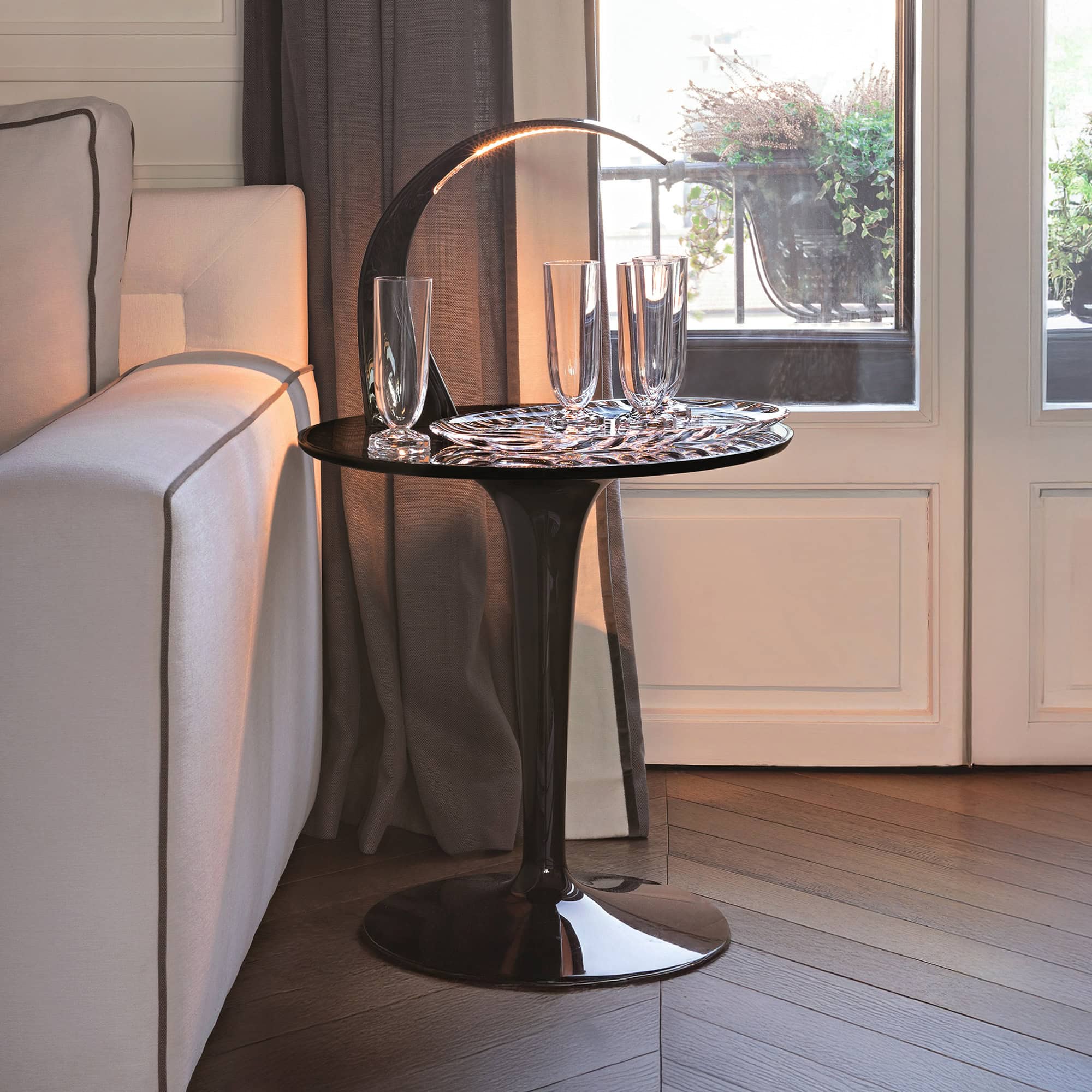 A modern, dark side table with an integrated curved LED lamp, holding three glass tumblers and a decorative tray. A light-colored sofa, dark curtains, and a bright window with balcony plants are in the background. Books are stacked on a dark wooden floor.