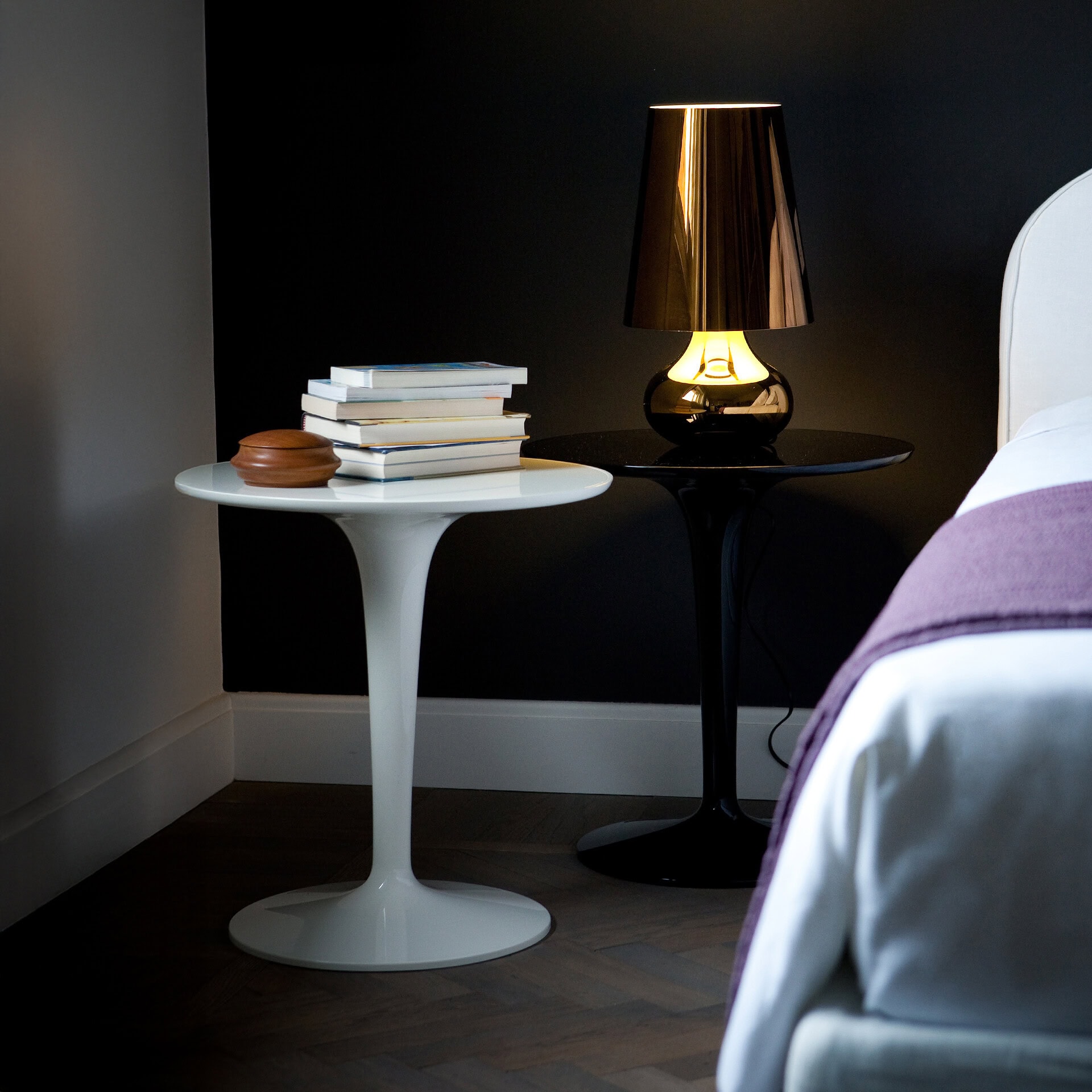 A modern bedroom scene with a white side table holding books and a wooden box, and a black side table with an illuminated bronze lamp. A bed with white bedding and a purple throw is partially visible on the right, against a dark wall.