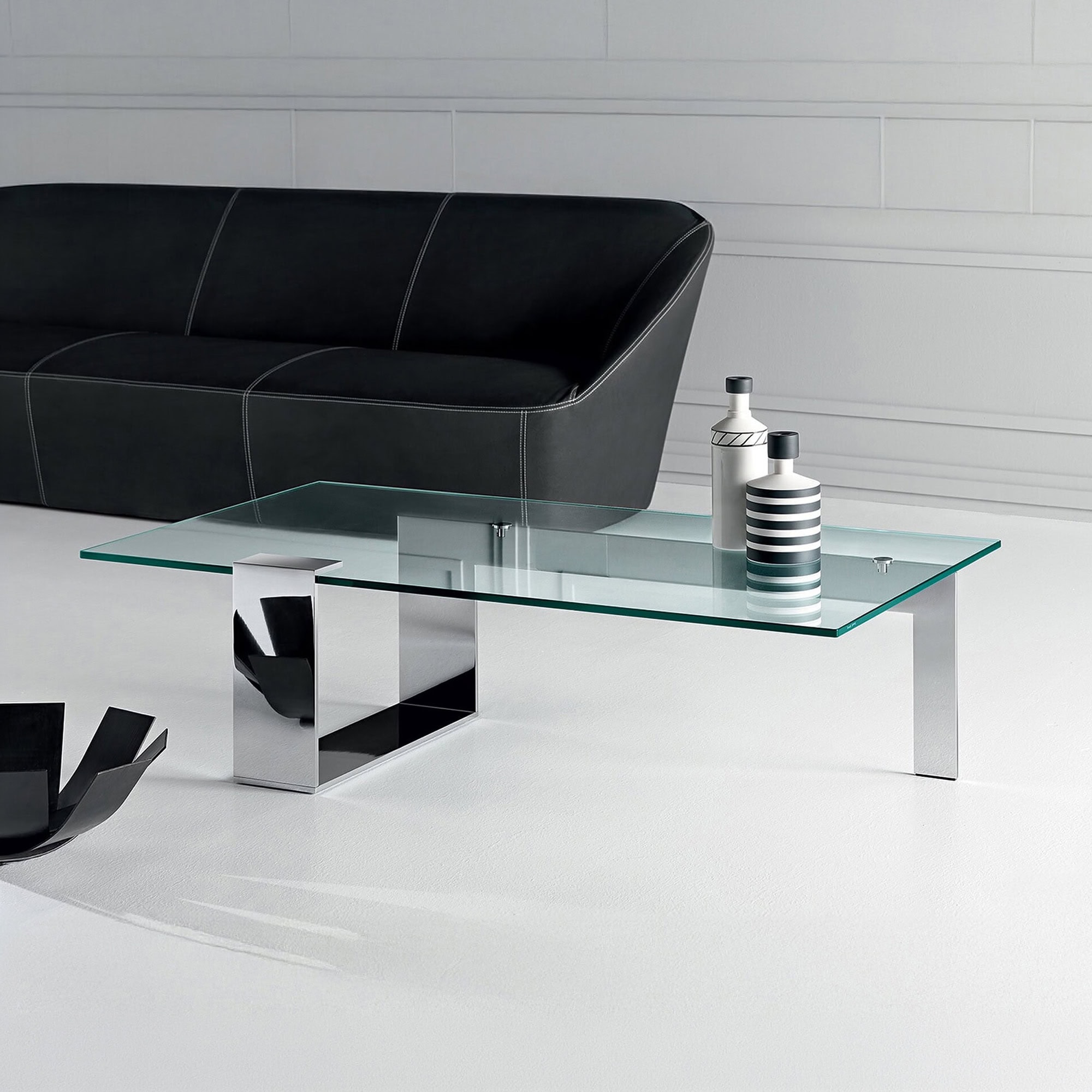 Plinsky Glass Low Coffee Table with glass top and black lacquered metal base placed in a contemporary living room with a dark sofa.