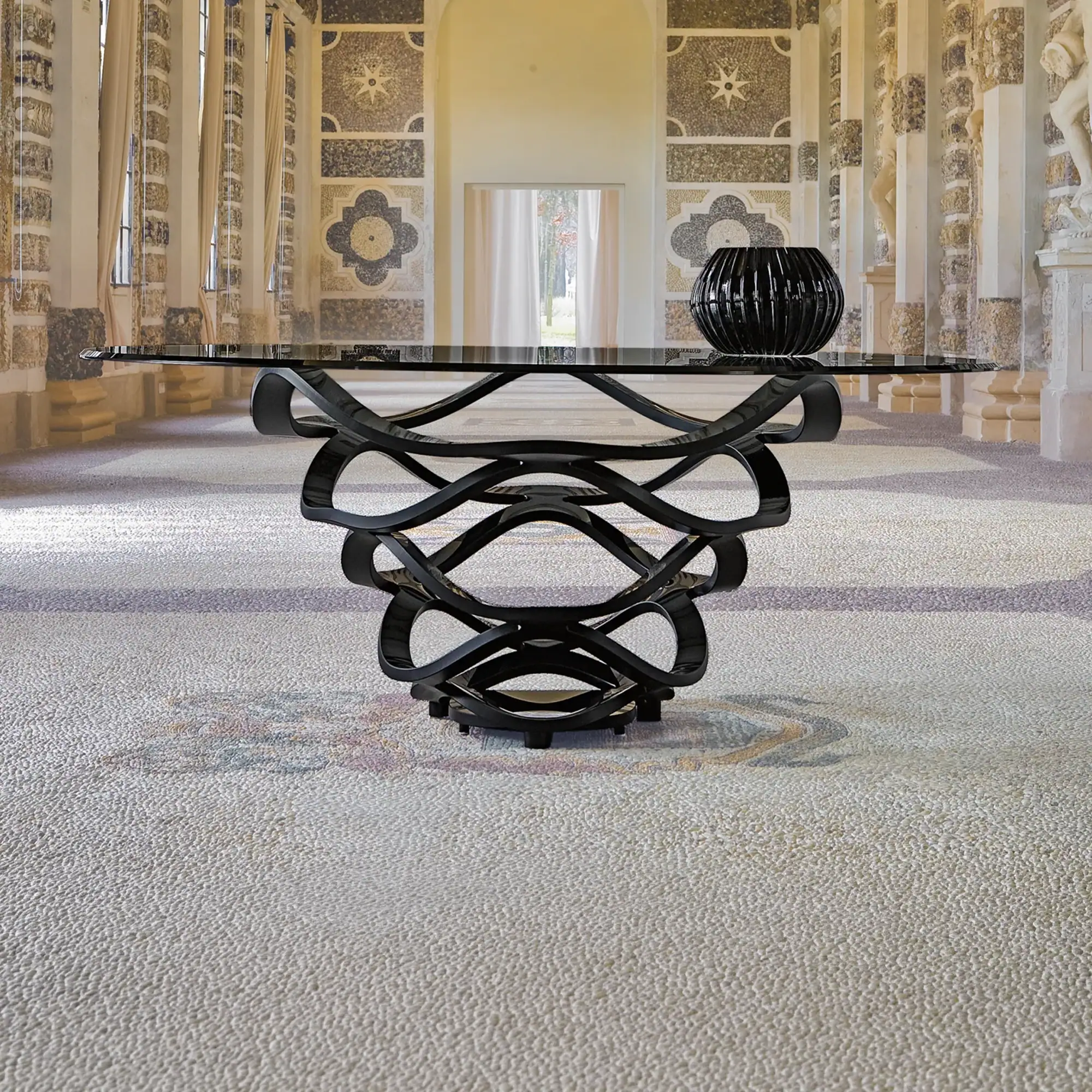A modern glass-top table with a black sculptural base in a grand hall with mosaic walls, arched windows, and classical statues.