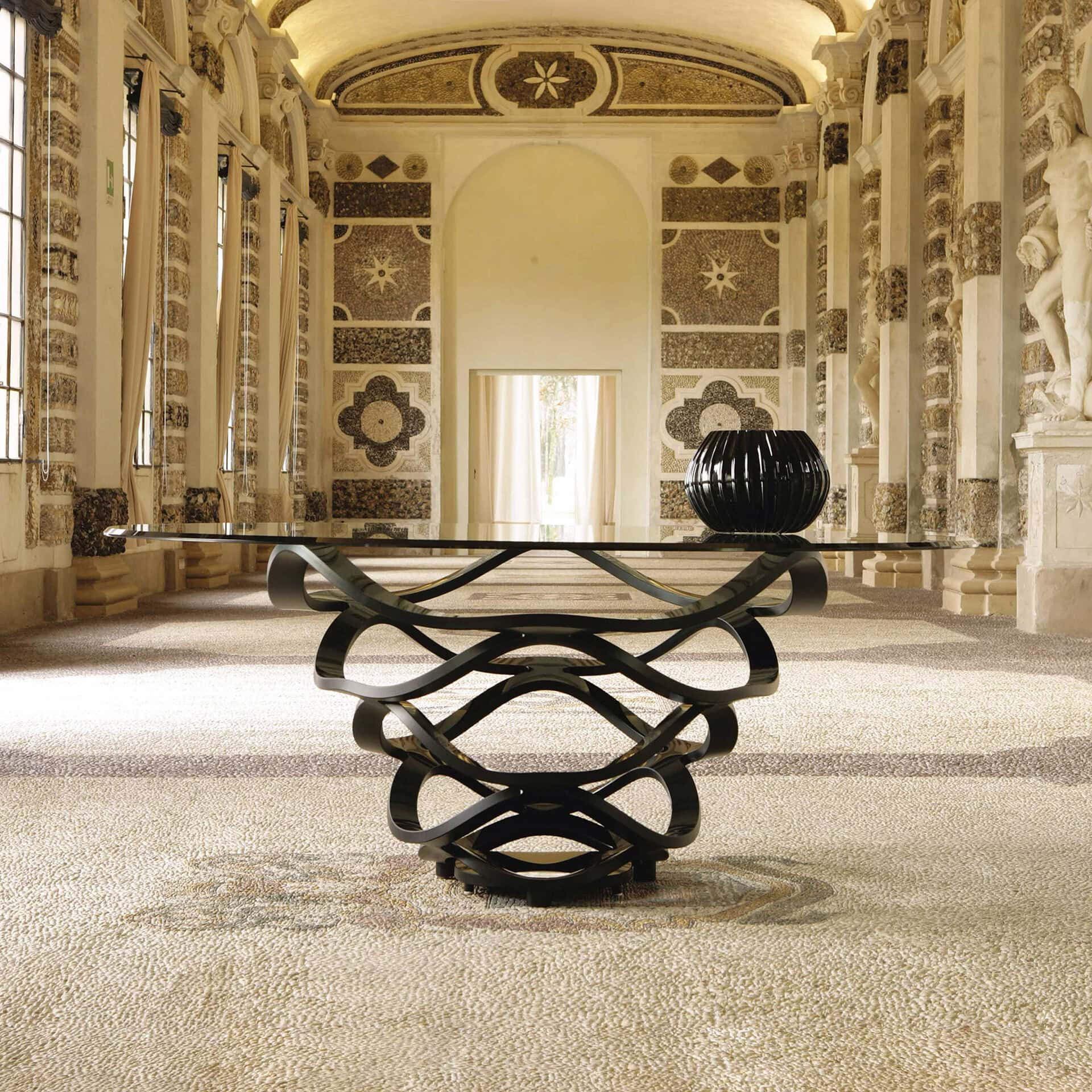 A modern glass-top table with a black sculptural base in a grand hall with mosaic walls, arched windows, and classical statues.