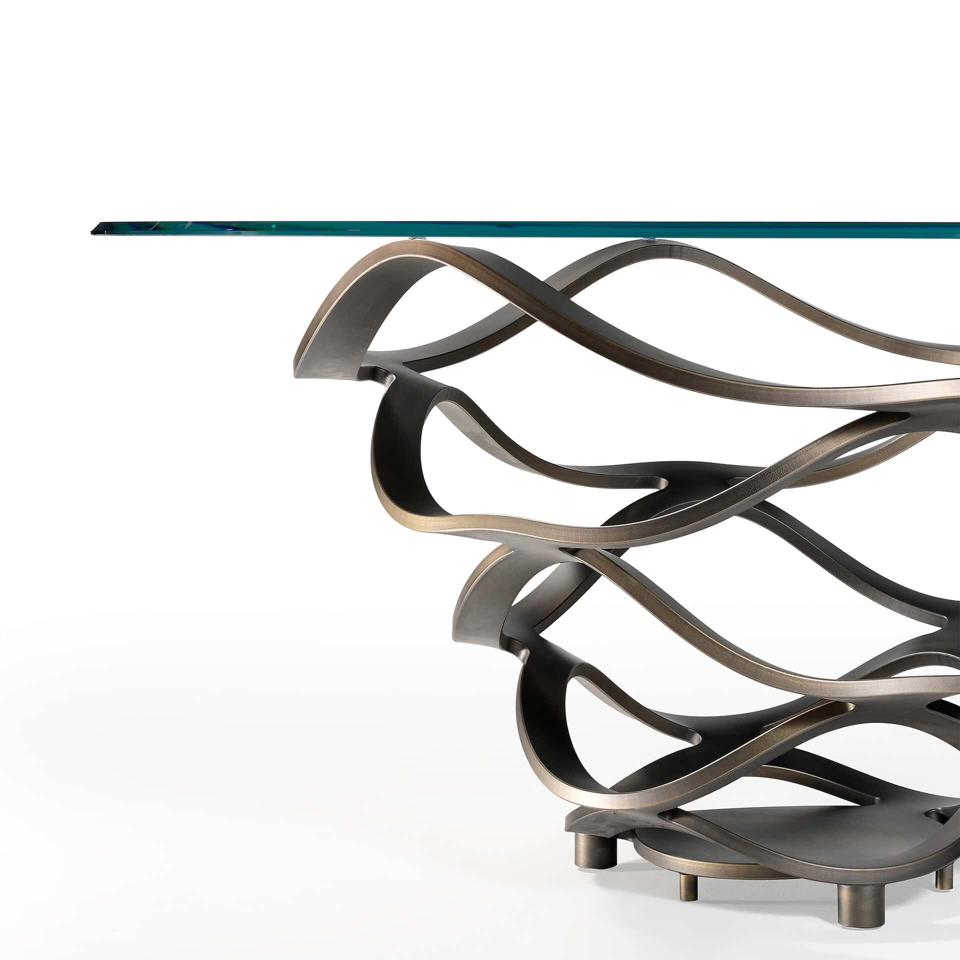 Close-up of a modern table with a sculptural, wavy bronze-toned metal base and a clear glass top, set against a white background.