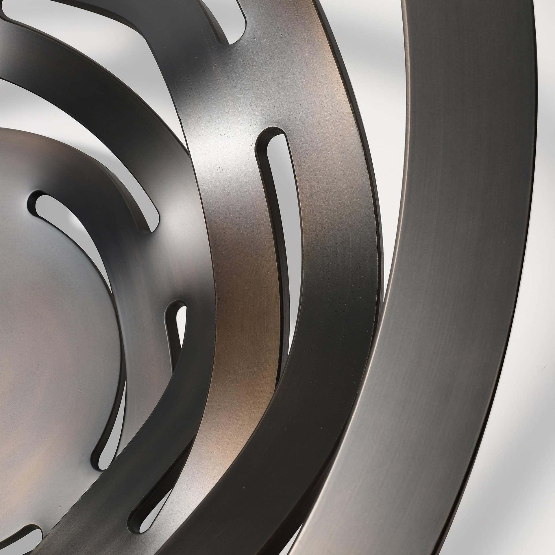 Close-up of an abstract sculpture made of dark, brushed metal curves and concentric bands with elongated cutouts, against a light background.