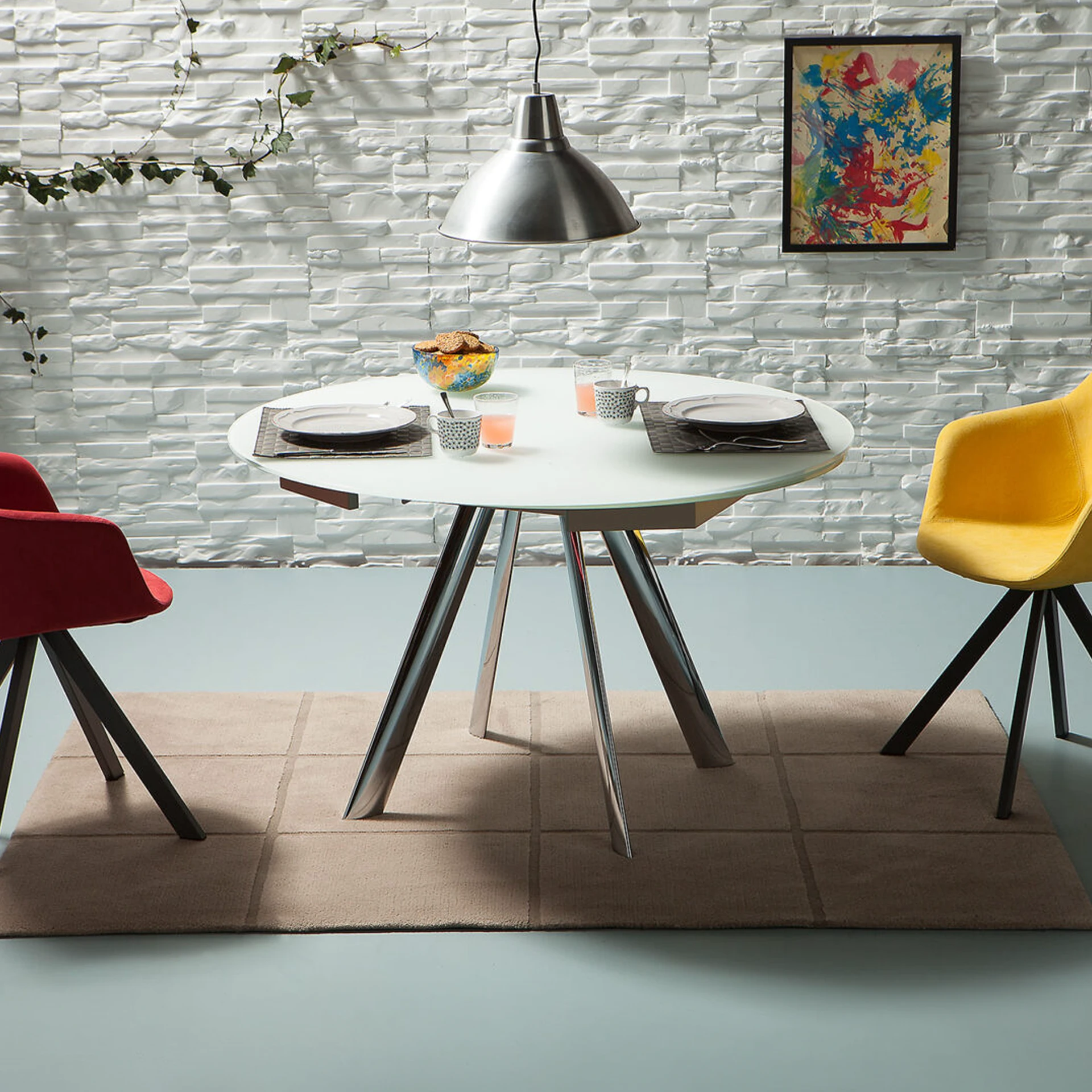 A modern dining room with a round white table, red and yellow chairs, a textured white brick wall, and a colorful abstract painting.