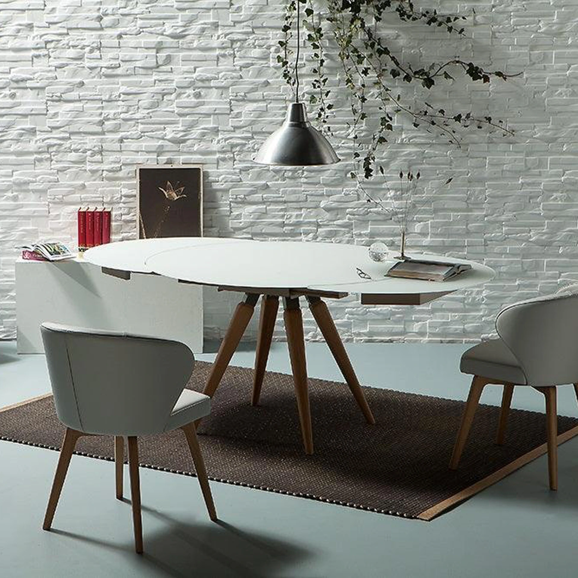 A modern dining room with a white oval table, gray chairs, and a white textured brick wall decorated with a green vine.
