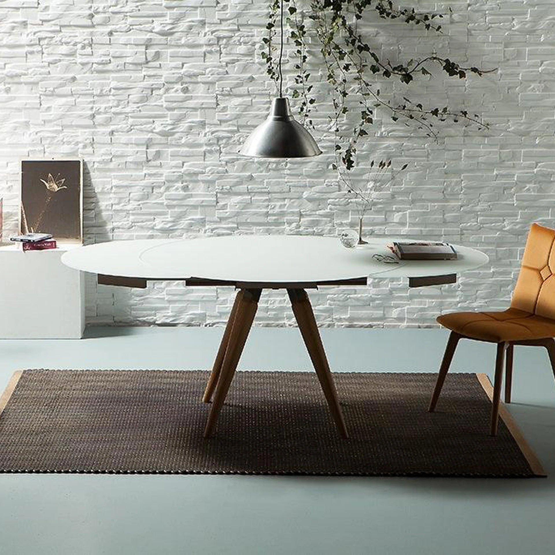 A modern dining room with a white oval table, a brown upholstered chair, and a white textured brick wall with a hanging vine.