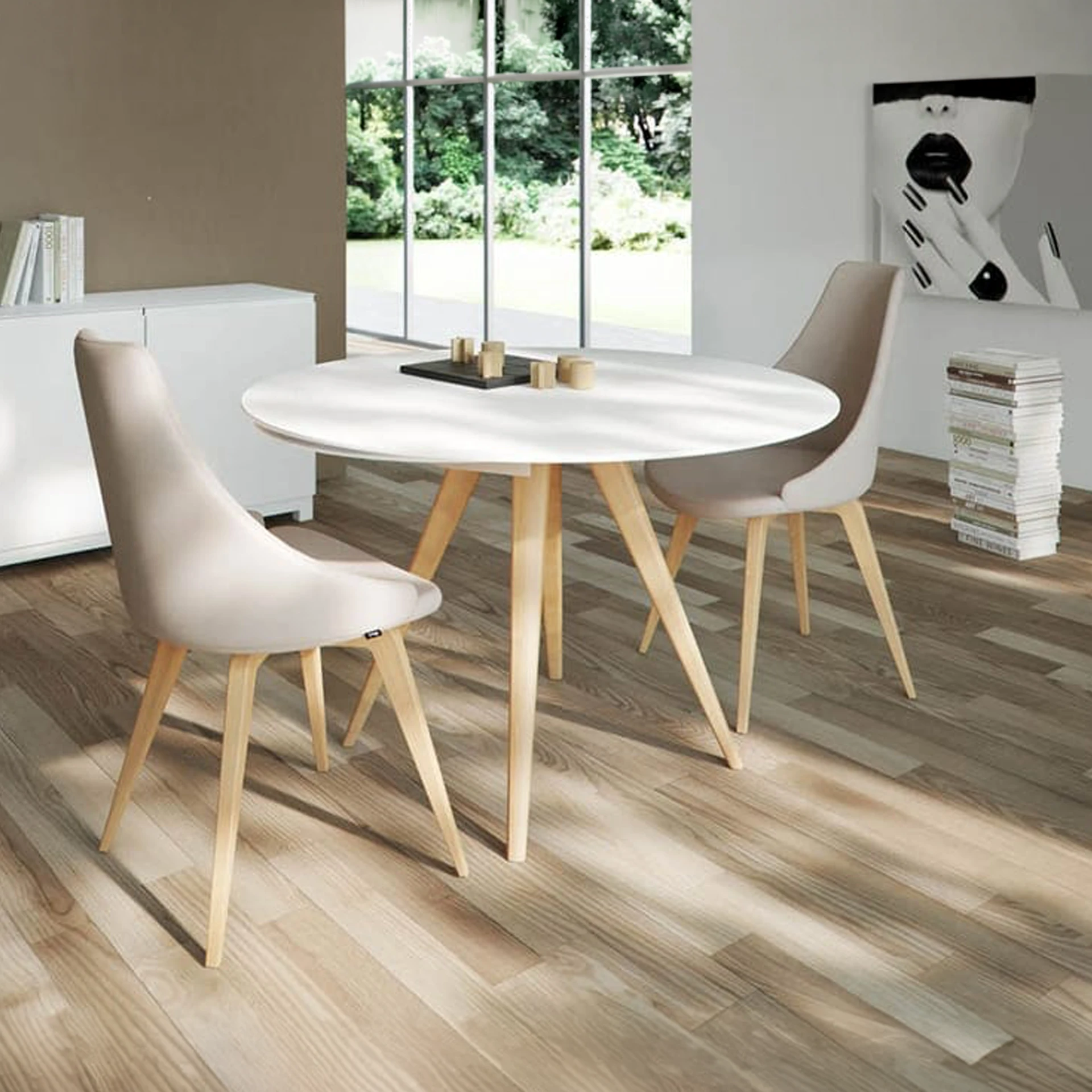 A modern dining room with a white round table, two beige chairs, and a large window overlooking green trees. A black and white abstract art piece hangs on the wall.