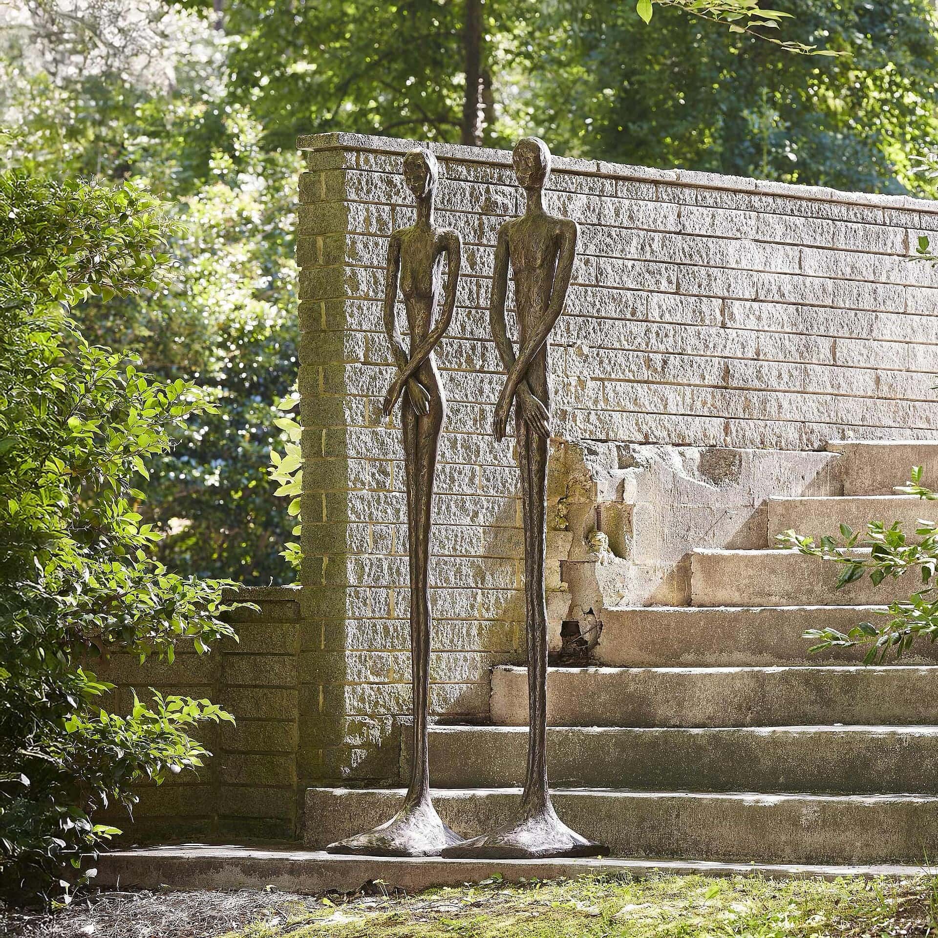 Artistic metal sculptures of tall, slender human forms outside a house in lush garden setting.