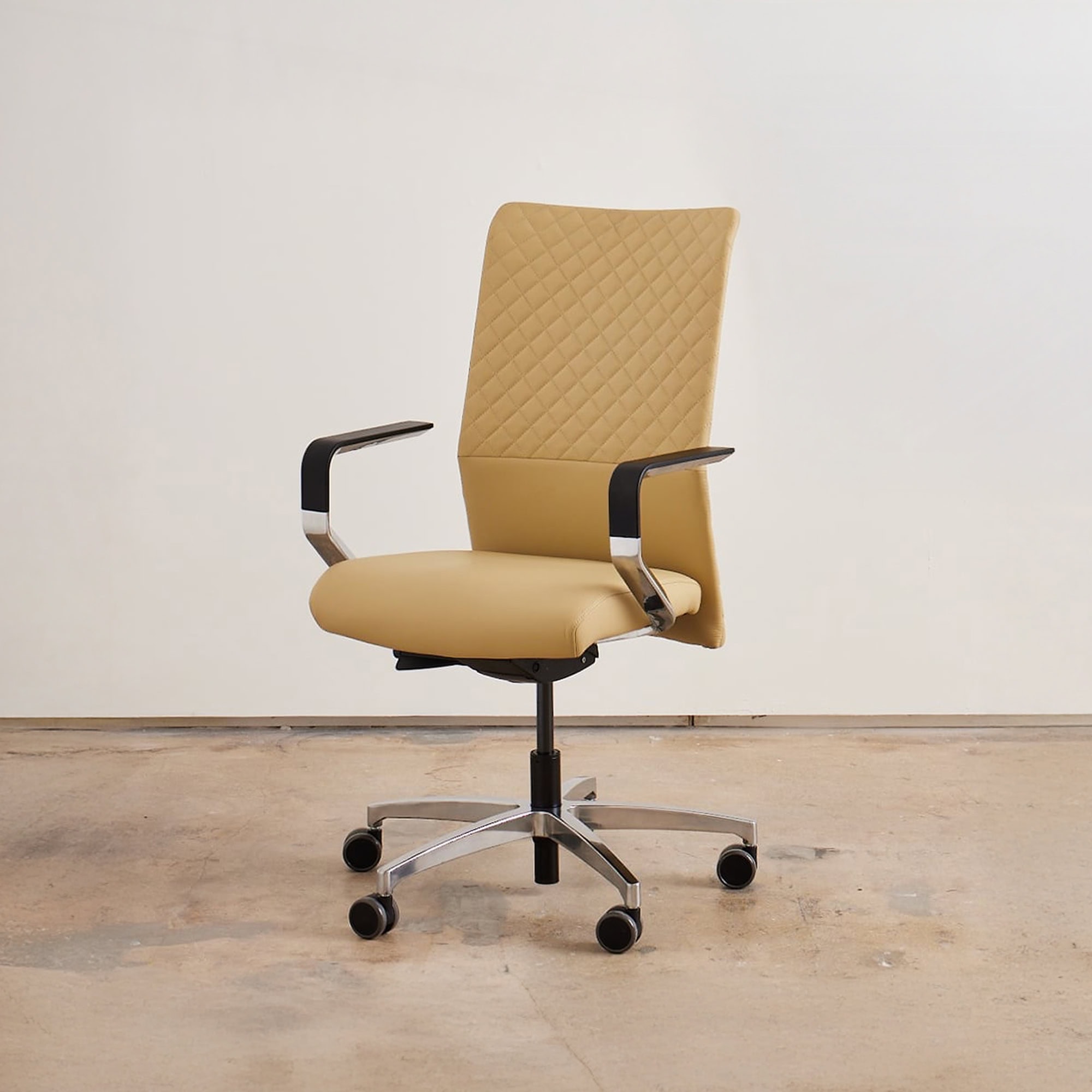 A modern tan office chair with a quilted back, chrome armrests, and a five-star wheeled base, set against a light wall and concrete floor.