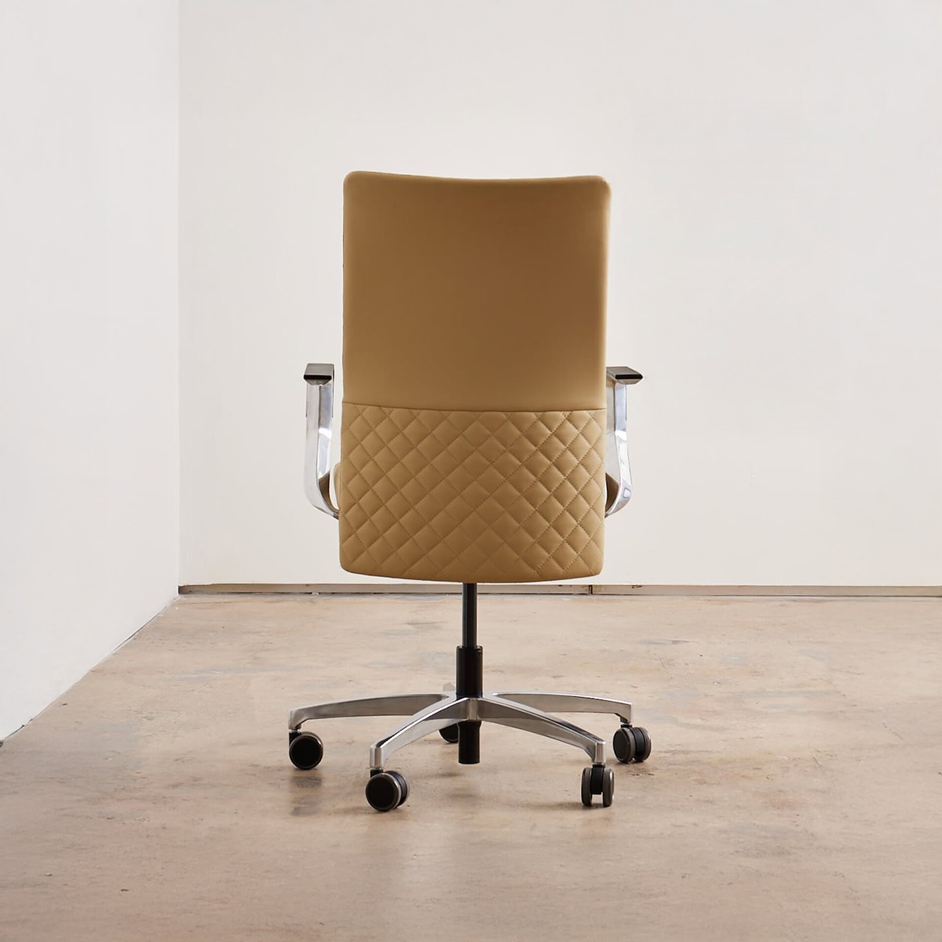 Rear view of a tan, high-backed office chair with a diamond-quilted lower back, polished chrome armrests, and a five-star wheeled base, set against a plain white wall and concrete floor.