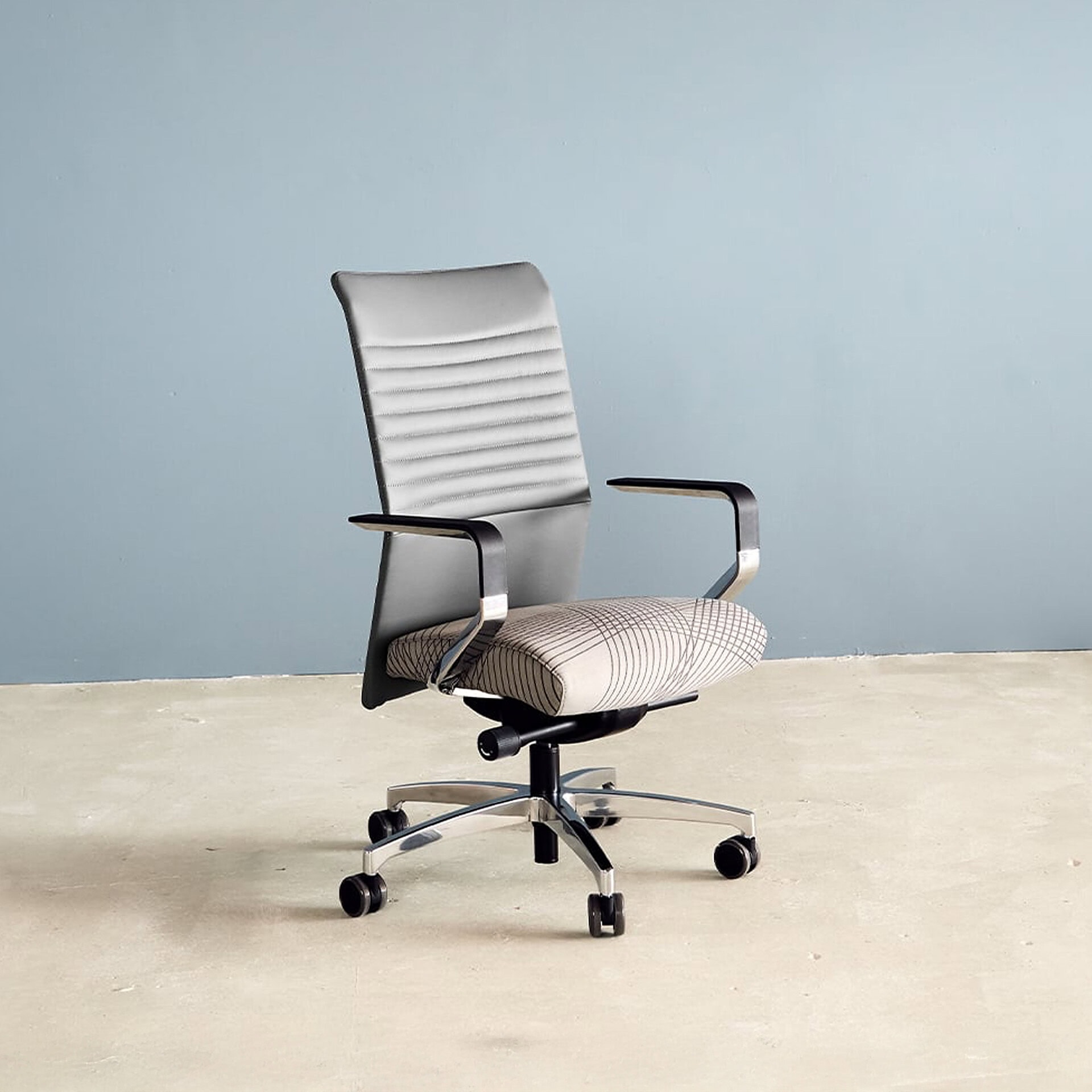 A modern office chair with a light gray backrest, a patterned beige seat, black armrests, and a polished metal five-star base on wheels, set against a light blue-gray wall.