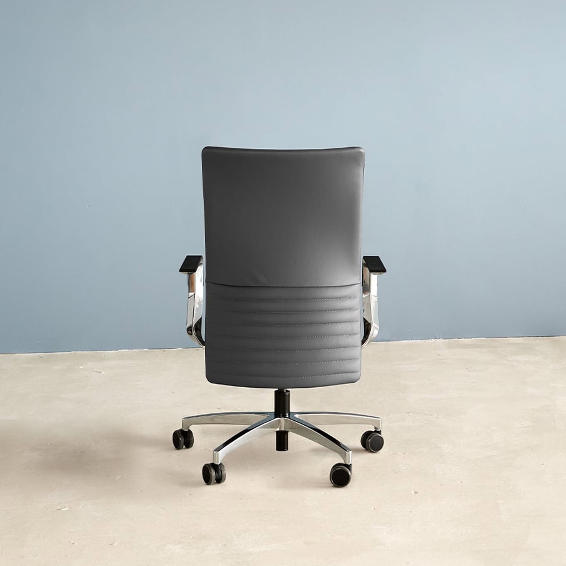 Rear view of a dark gray modern office chair with chrome armrests and a five-star wheeled base against a light blue wall and concrete floor.