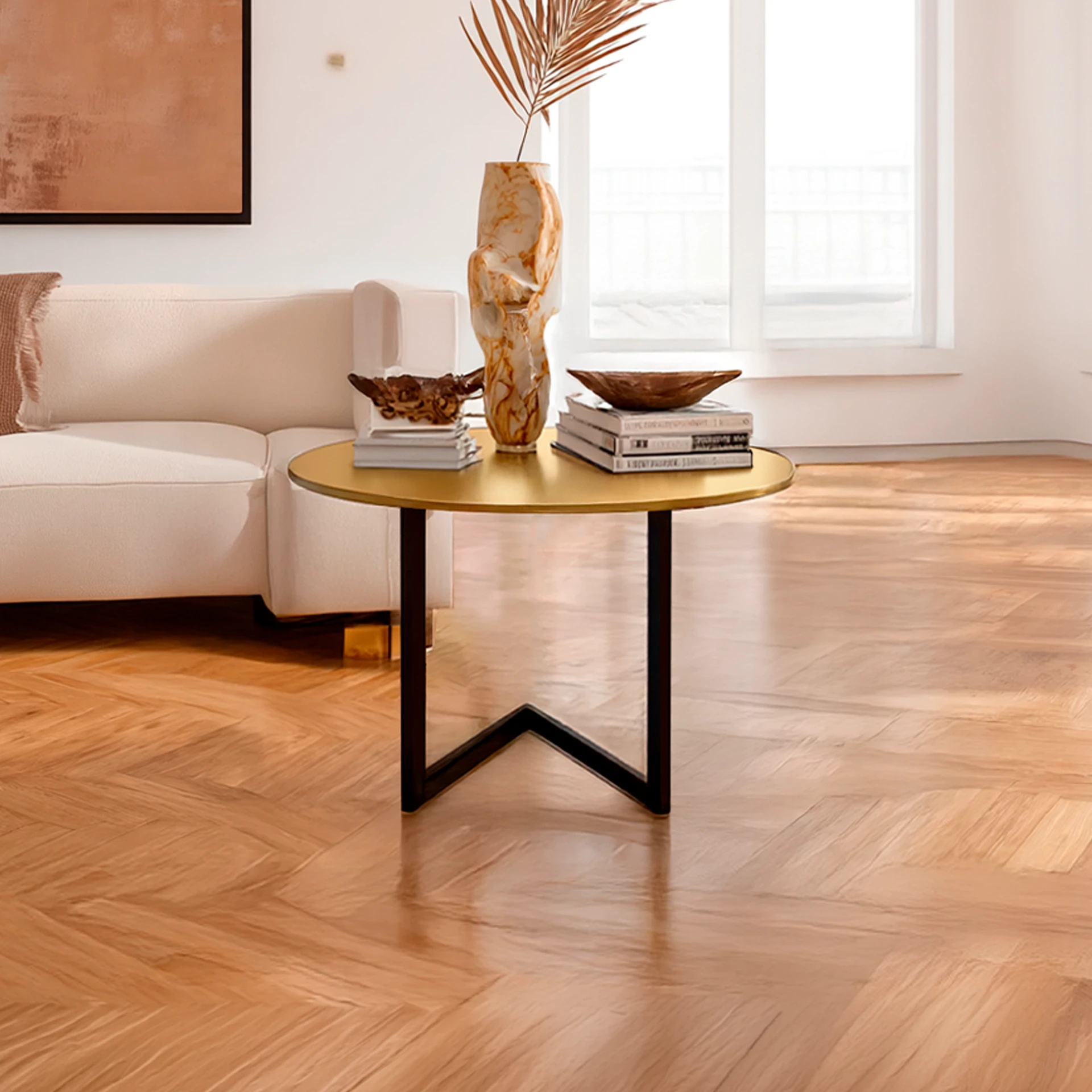 A modern living room with a gold coffee table, cream sofa, and parquet flooring. The table holds a marbled vase with a dried palm frond, books, and decorative bowls. A large window provides natural light.