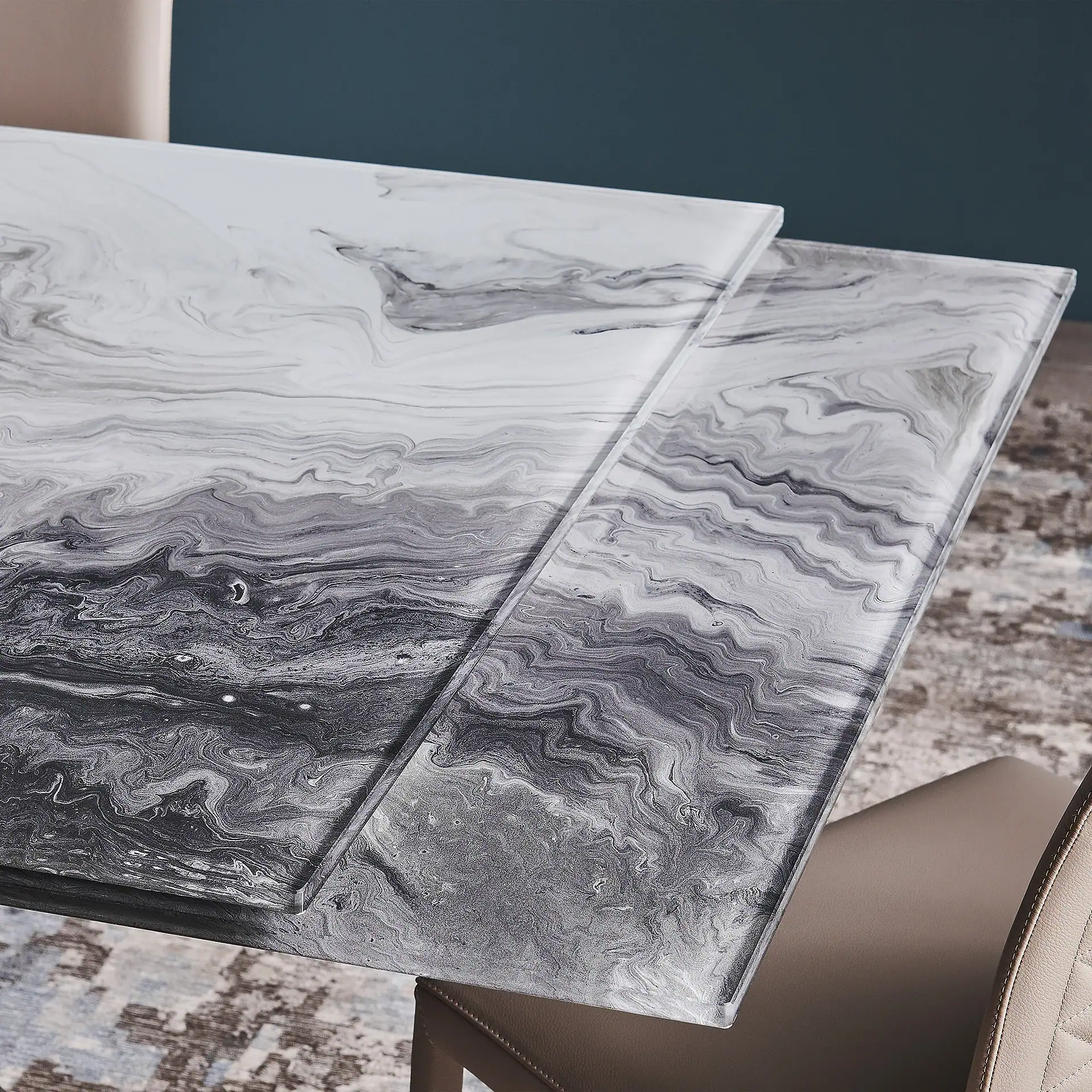 Close-up of a modern dining table with a white, gray, and black marbled glass top, showing two offset glass layers. A beige chair, patterned rug, and dark wall are in the background.