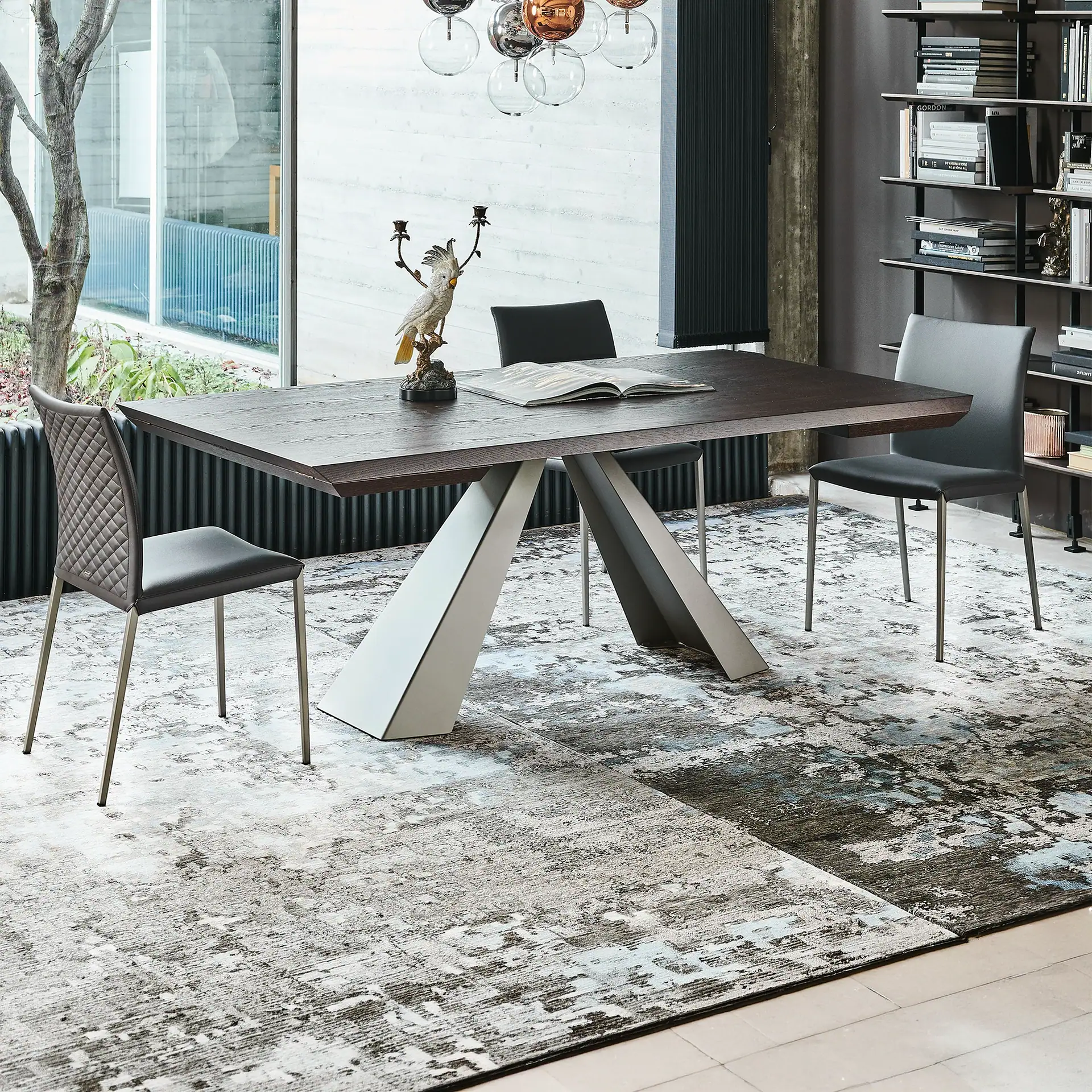 A modern dining room with a dark wood table, geometric base, four gray chairs, abstract rug, cockatoo figurine, and a spherical chandelier.