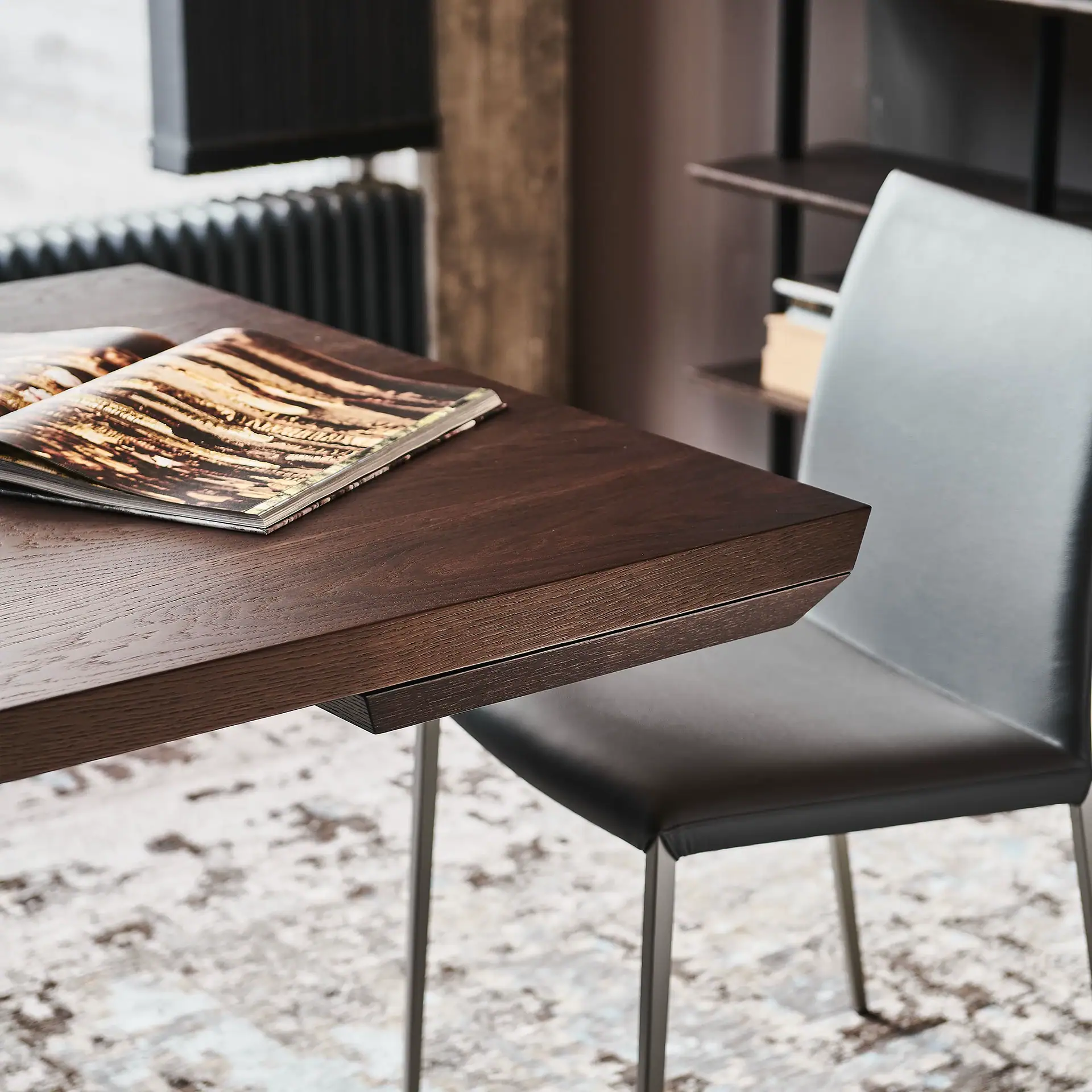 A dark wooden table with an open magazine on it, next to a modern dark gray chair. A patterned rug is on the floor, and a window and bookshelf are in the background.