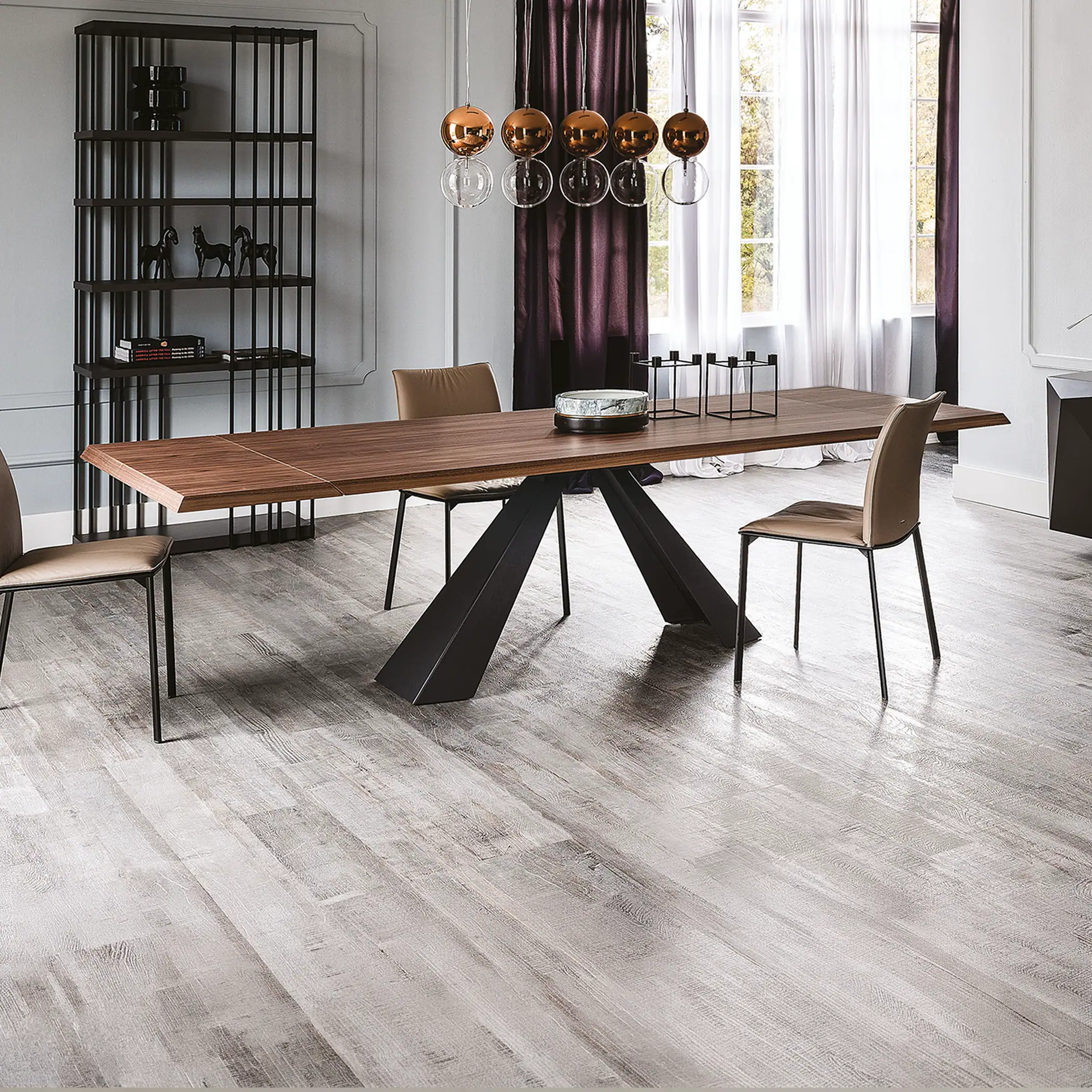 A modern dining room with a long wooden table, light brown chairs, a unique chandelier, and a tall black bookshelf against light grey walls and distressed wood floors.
