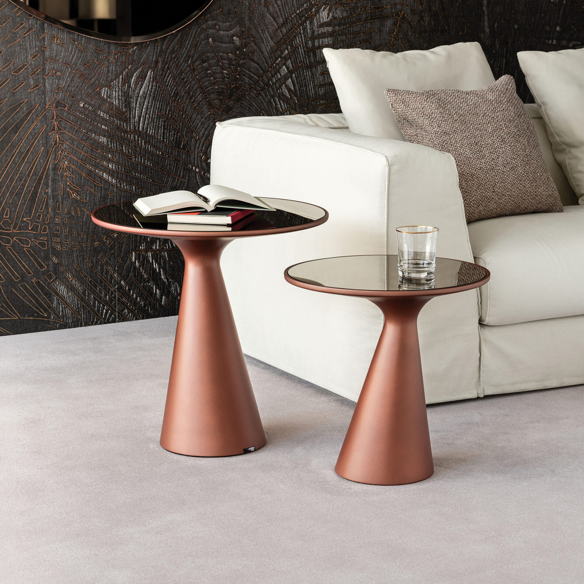Two modern side tables with copper bases and dark glass tops next to an off-white sofa. The taller table has books, and the shorter one holds a glass. A dark wall with metallic palm leaf patterns is in the background.
