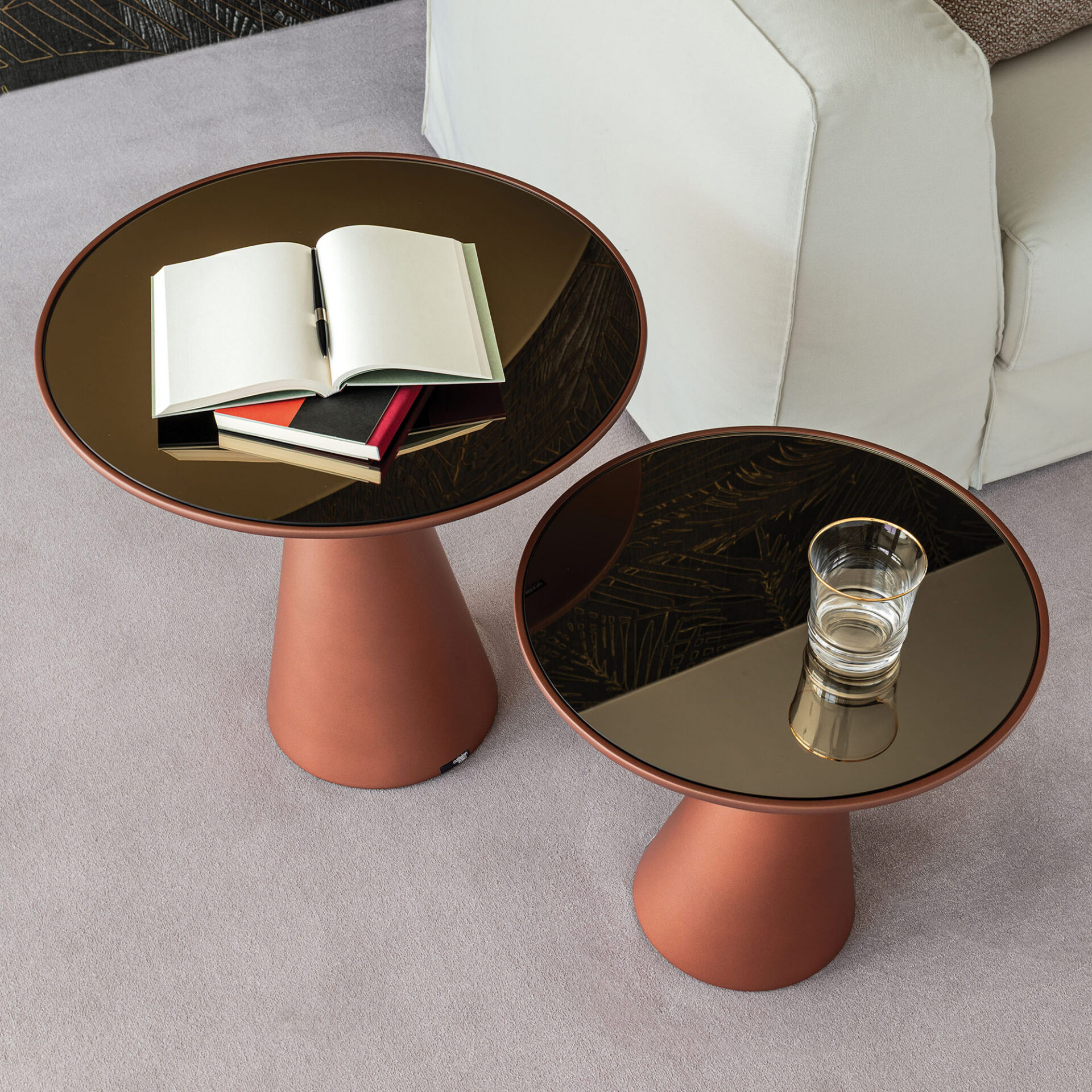 Two modern round side tables with terracotta bases and dark bronze glass tops on a light grey carpet. The larger table holds an open notebook and pen, while the smaller one has a glass of water. A light sofa is in the background.