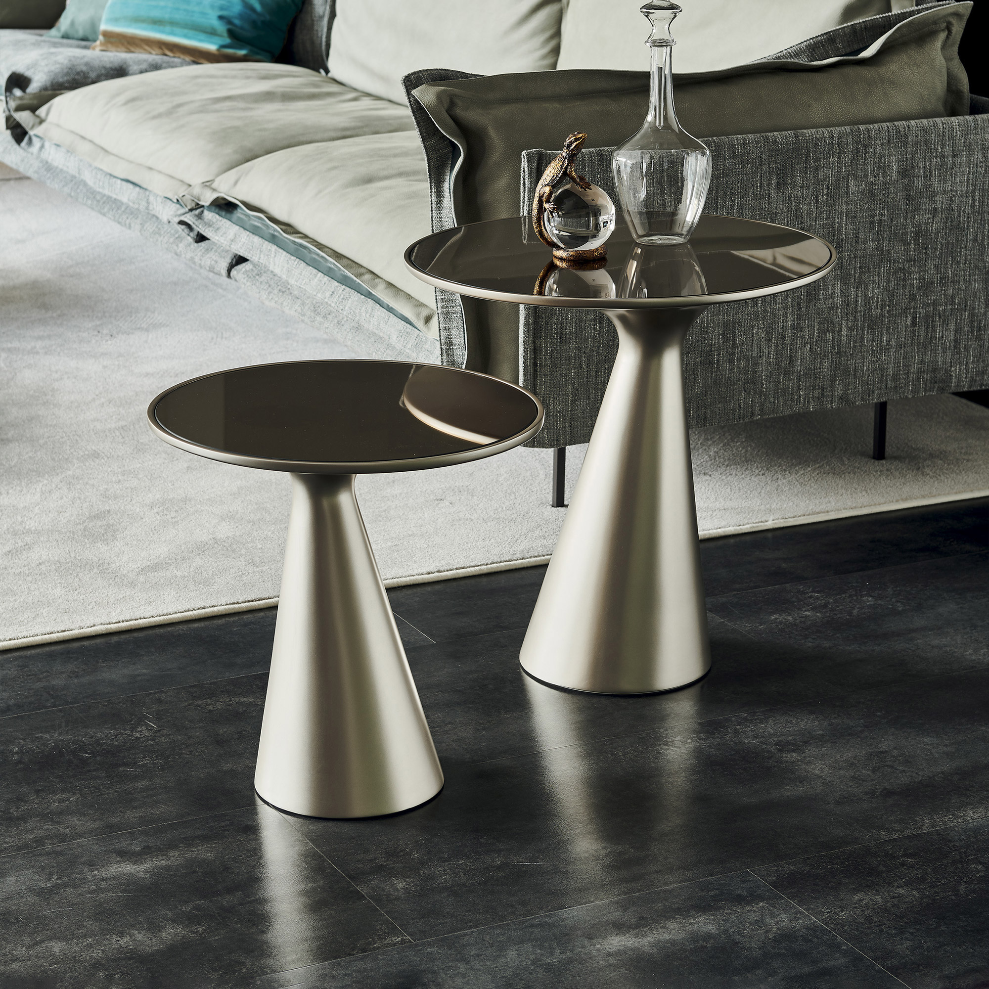 Two modern, conical-based side tables with dark, reflective tops in a living room. A glass decanter and a lizard figurine adorn the taller table. A grey sofa with a teal pillow is in the background.