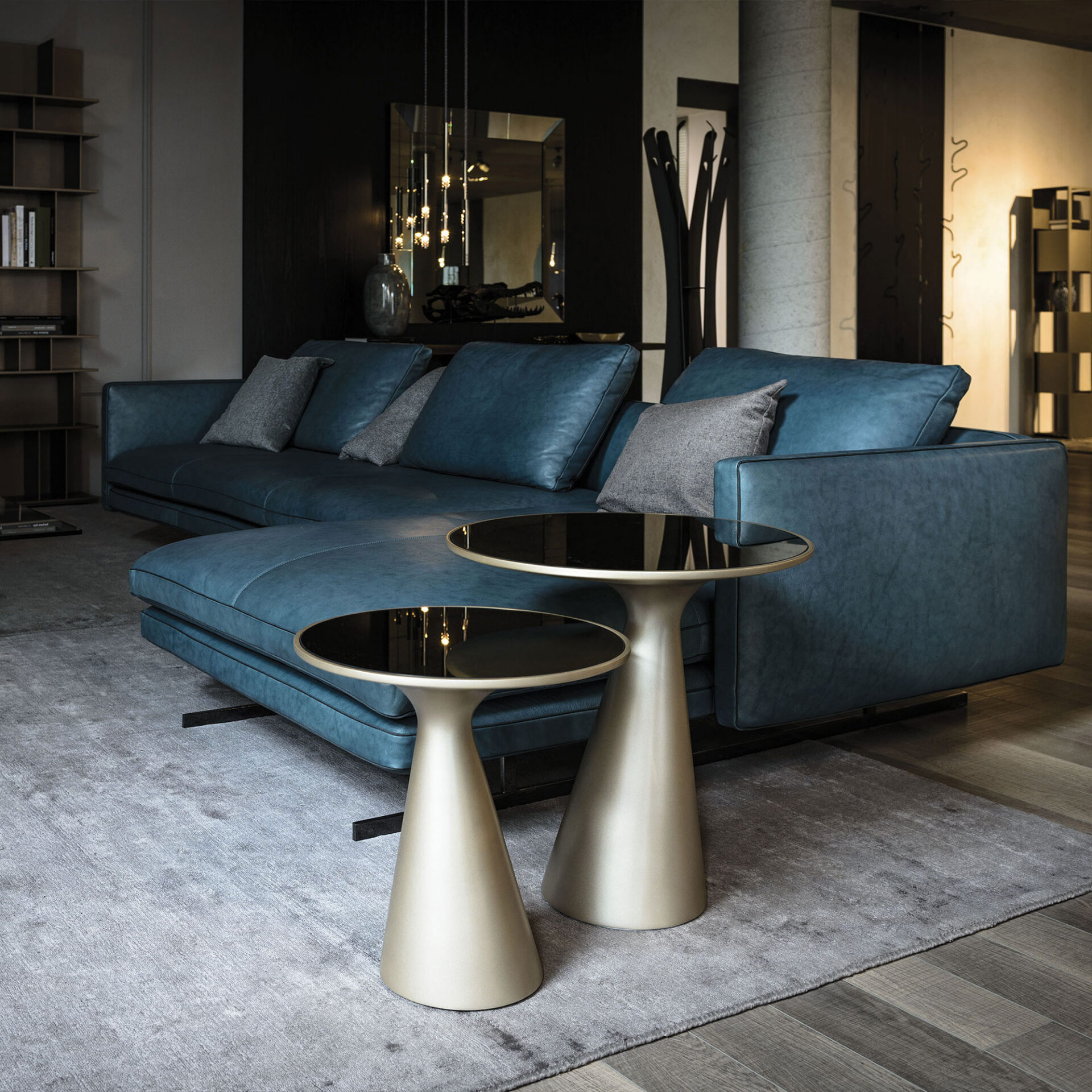 A modern living room featuring a large, deep teal leather sectional sofa with gray throw pillows. Two champagne-colored side tables with black glass tops sit on a gray rug. The room has dark walls, a large mirror reflecting pendant lights, and various contemporary decor elements.