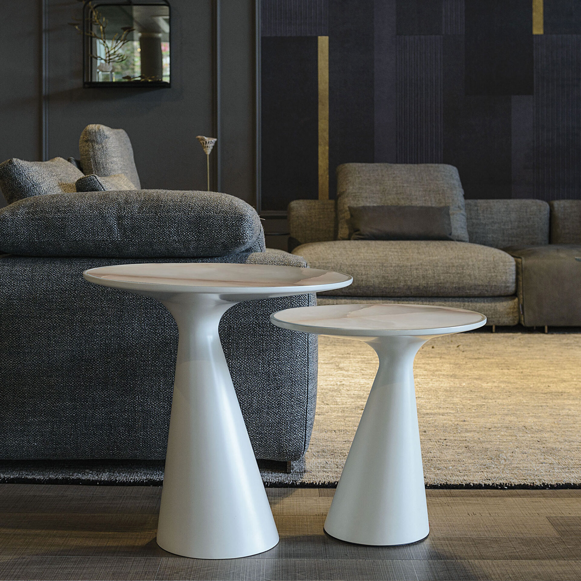 Two white, sculptural side tables with marble-patterned tops in front of a dark grey textured sectional sofa. The room has dark walls with geometric patterns and a light-colored rug.