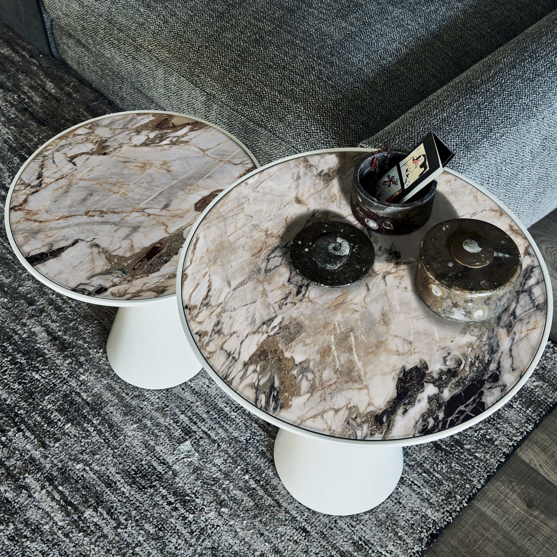 Two round marble-topped side tables with white bases, placed on a grey rug next to a grey sofa. The marble tops have prominent dark veining, and one table holds decorative objects.