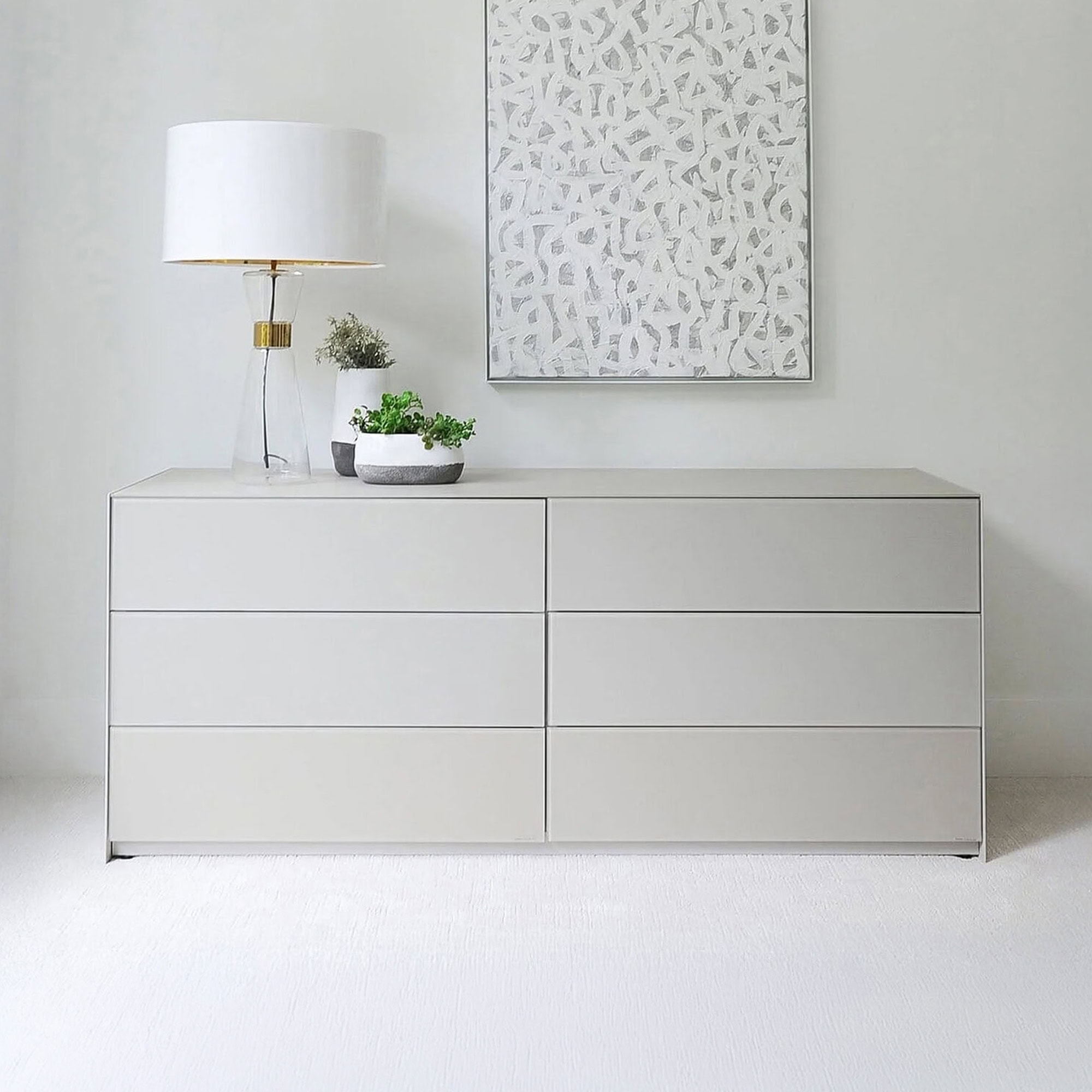 A modern light grey dresser with six drawers, topped with a glass lamp and two potted plants. An abstract white and grey artwork hangs above it on a light grey wall, with a light-colored carpet below.