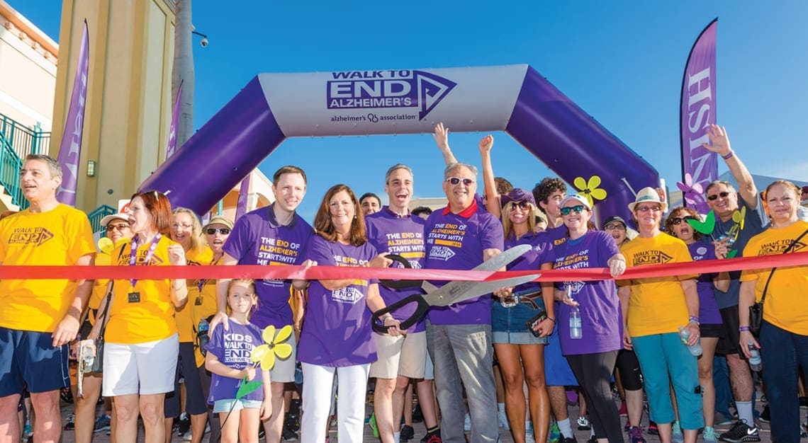 Alzheimer's awareness event with a crowd holding a purple arch and ribbon.