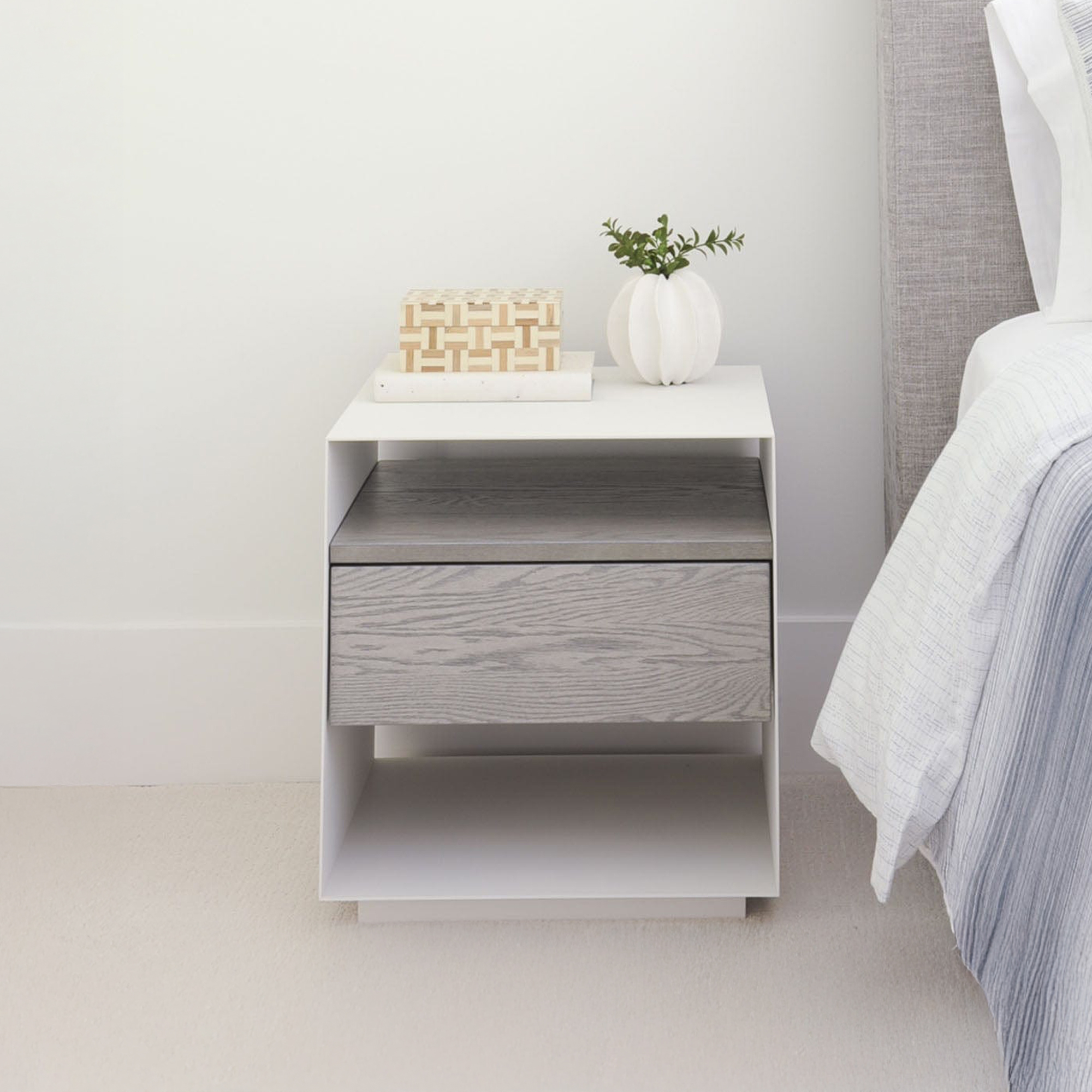 A modern white nightstand with a grey wood-grain drawer, topped with a decorative box and a white vase with greenery, next to a bed with a grey headboard and light blue bedding.