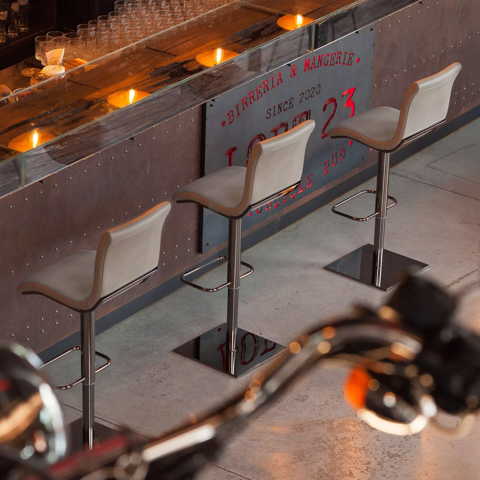 A modern bar interior with three light beige bar stools, a dark wood and glass bar top with warm lights, and a riveted metal bar front with red text reading 'MOTORCYCLE PUB.' Motorcycle handlebars are blurred in the foreground.
