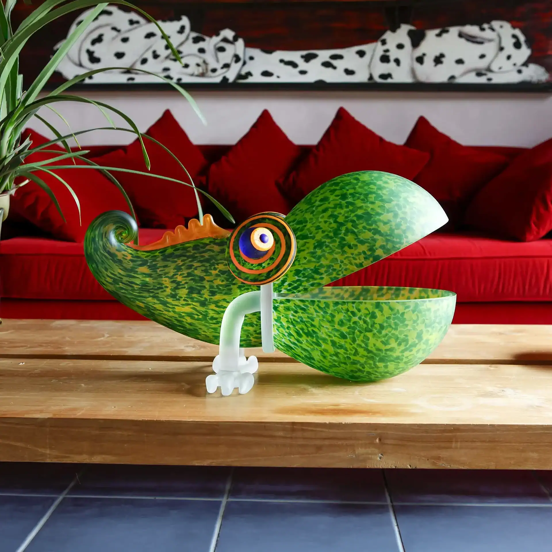 Chameleon Big Glass Bowl with an open mouth and spiral eye sits on a wooden table in front of a red sofa and a painting of a Dalmatian.