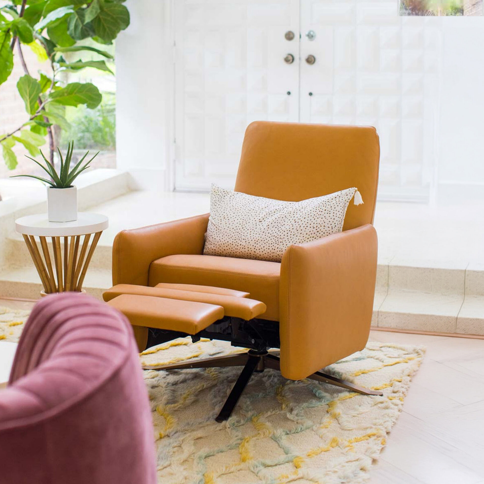 A modern living room with a tan leather recliner, white polka dot pillow, patterned shag rug, and a side table with a plant. A pink velvet chair is partially visible in the foreground.