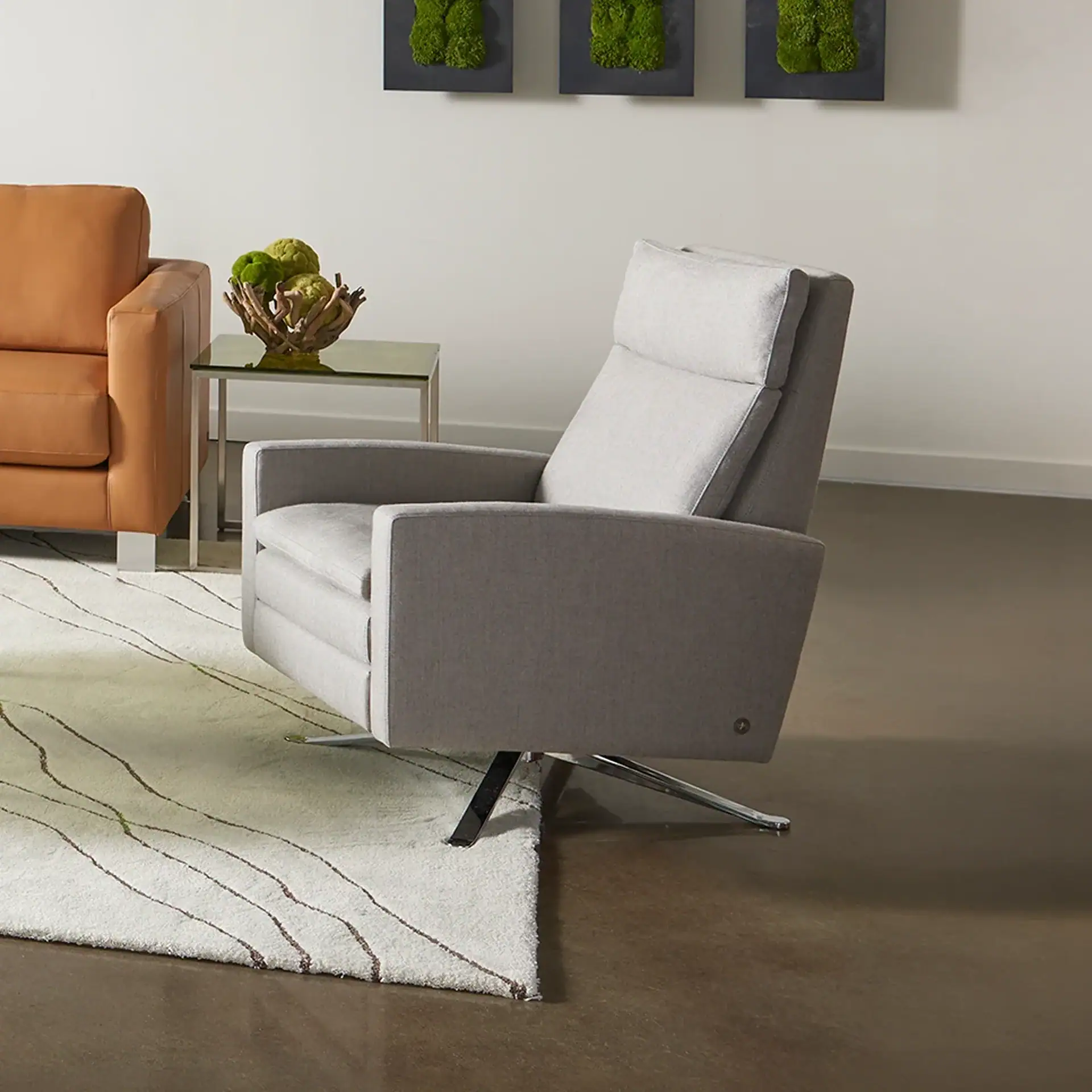 A light gray upholstered armchair with a chrome swivel base in a modern living room. A tan leather sofa, glass side table with moss decor, and wall art are also visible.