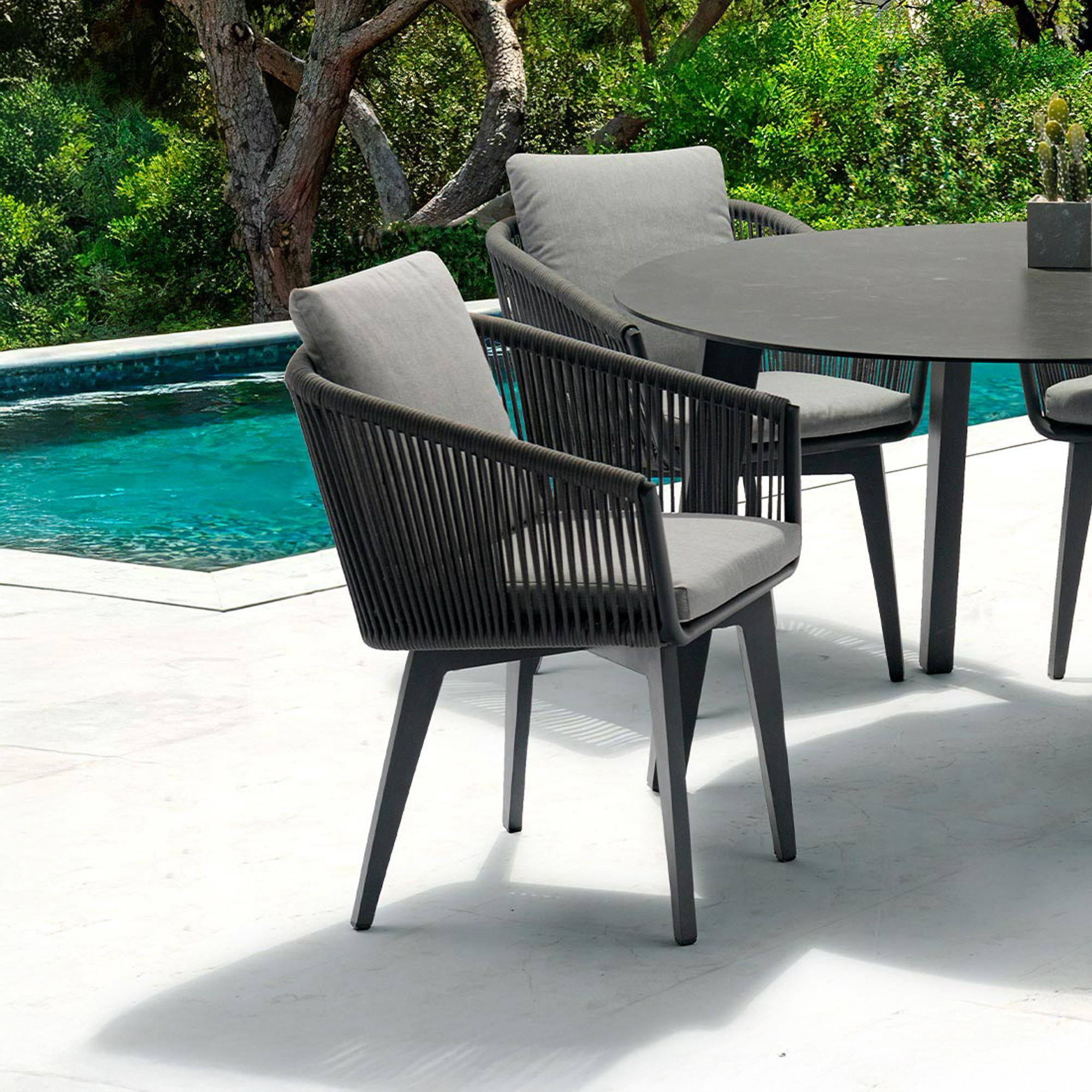 Modern dark gray outdoor dining set with light cushions on a white patio next to a turquoise swimming pool, surrounded by lush green trees.