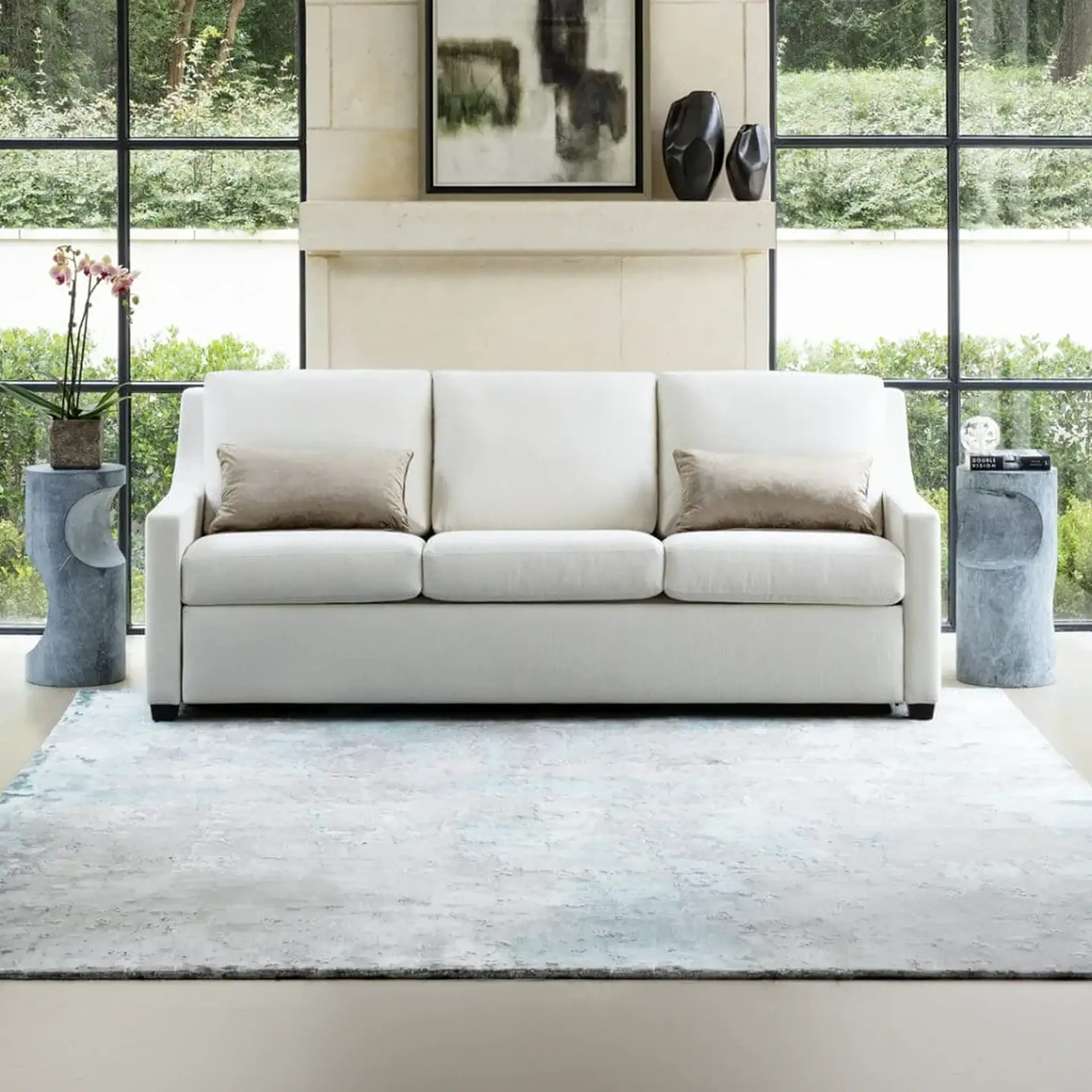 A white sofa with two taupe pillows sits on a light blue and gray rug in front of large windows overlooking green foliage. A stone mantel above the sofa displays abstract art and dark vases. Two textured gray side tables flank the sofa, holding plants, books, and decorative items.