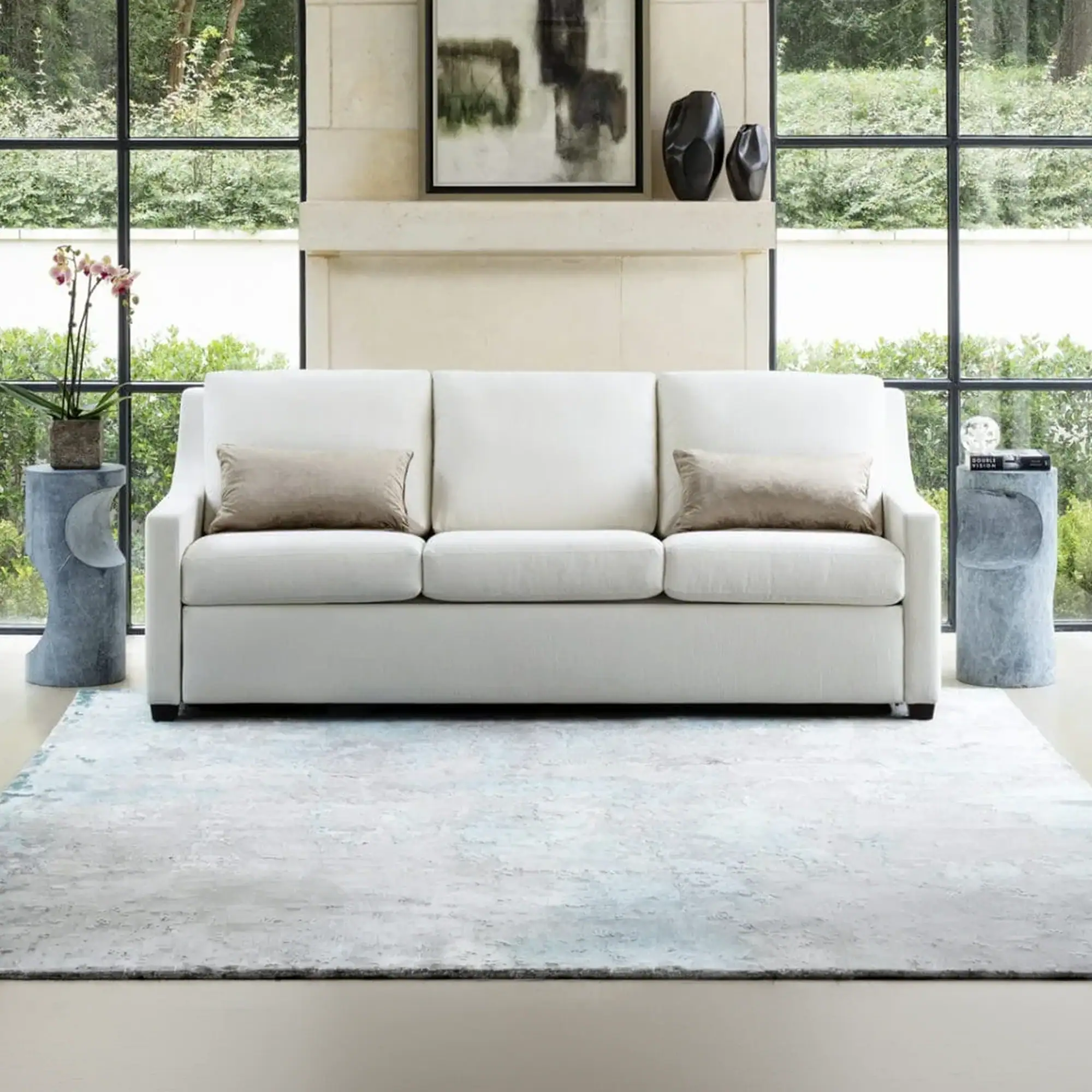 A white sofa with two taupe pillows sits on a light blue and gray rug in front of large windows overlooking green foliage. A stone mantel above the sofa displays abstract art and dark vases. Two textured gray side tables flank the sofa, holding plants, books, and decorative items.