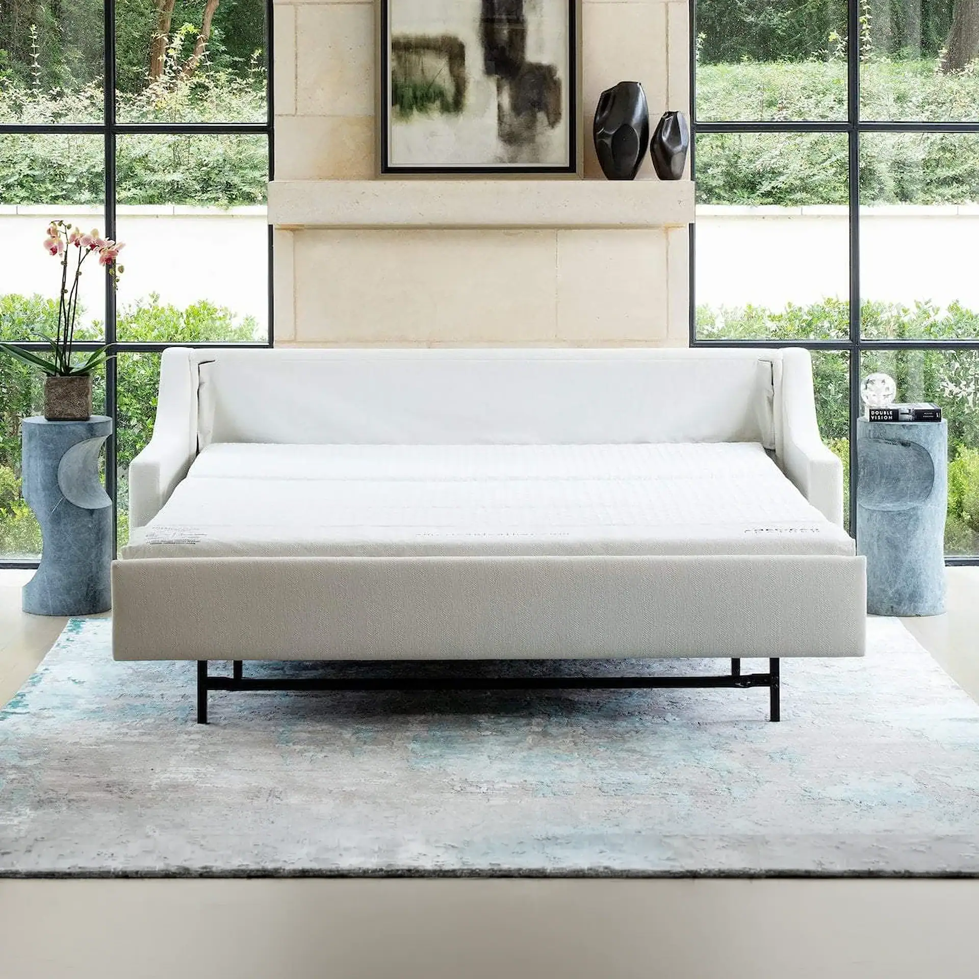 A modern light-colored sofa bed in a bright room with large windows overlooking greenery. The room features a patterned rug, stone wall, abstract art, and decorative side tables.