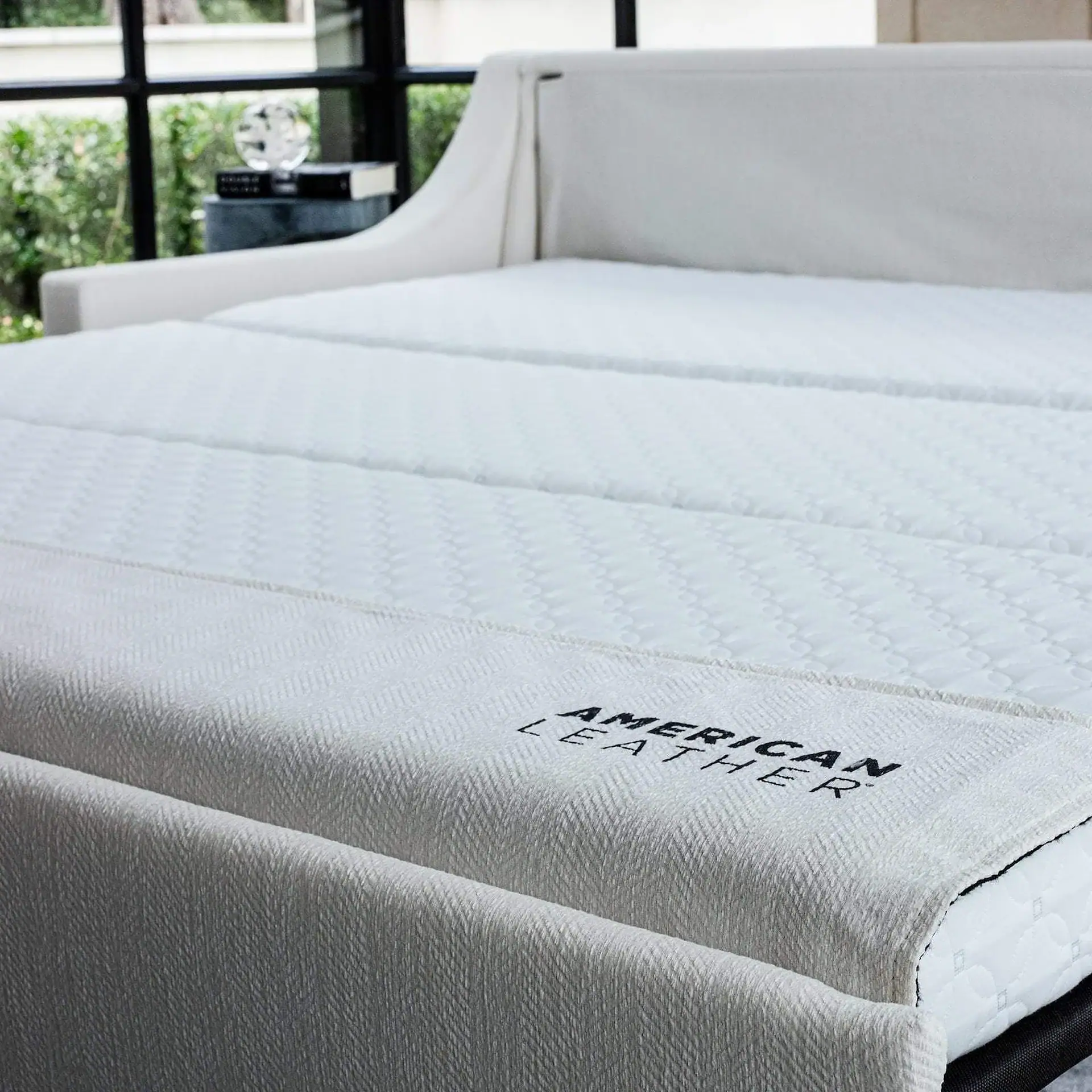 A white bed with a quilted mattress and a white blanket embroidered with "AMERICAN LEATHER" in a bright room with a window showing green foliage.