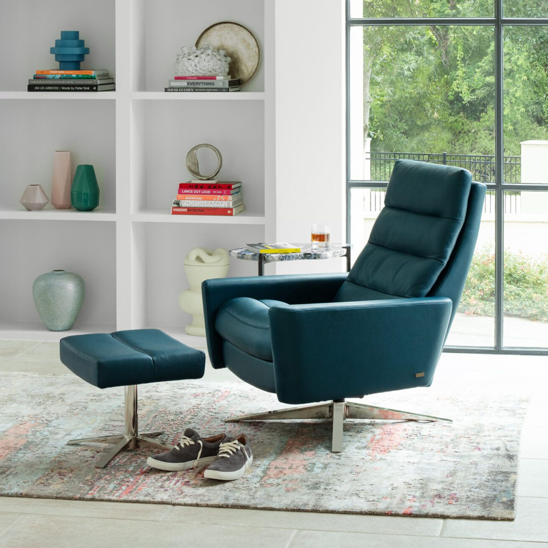 A modern teal blue recliner and matching ottoman in a bright room. The chair faces a large window with a view of green trees. A white bookshelf with decorative items and books is on the left, and a small side table with a drink is on the right. A pair of sneakers rests on an abstract rug in the foreground.