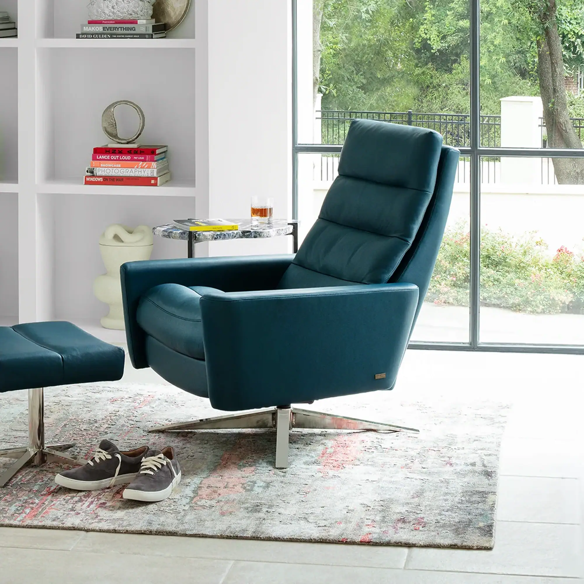 A modern teal blue recliner and matching ottoman in a bright room. The chair faces a large window with a view of green trees. A white bookshelf with decorative items and books is on the left, and a small side table with a drink is on the right. A pair of sneakers rests on an abstract rug in the foreground.