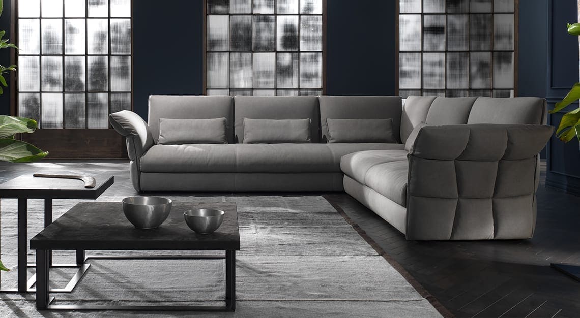 A stylish L-shaped gray sectional sofa in a modern living room with dark walls, large frosted windows, and two dark coffee tables with metal bowls and a decorative object. A large green plant is visible on the left.