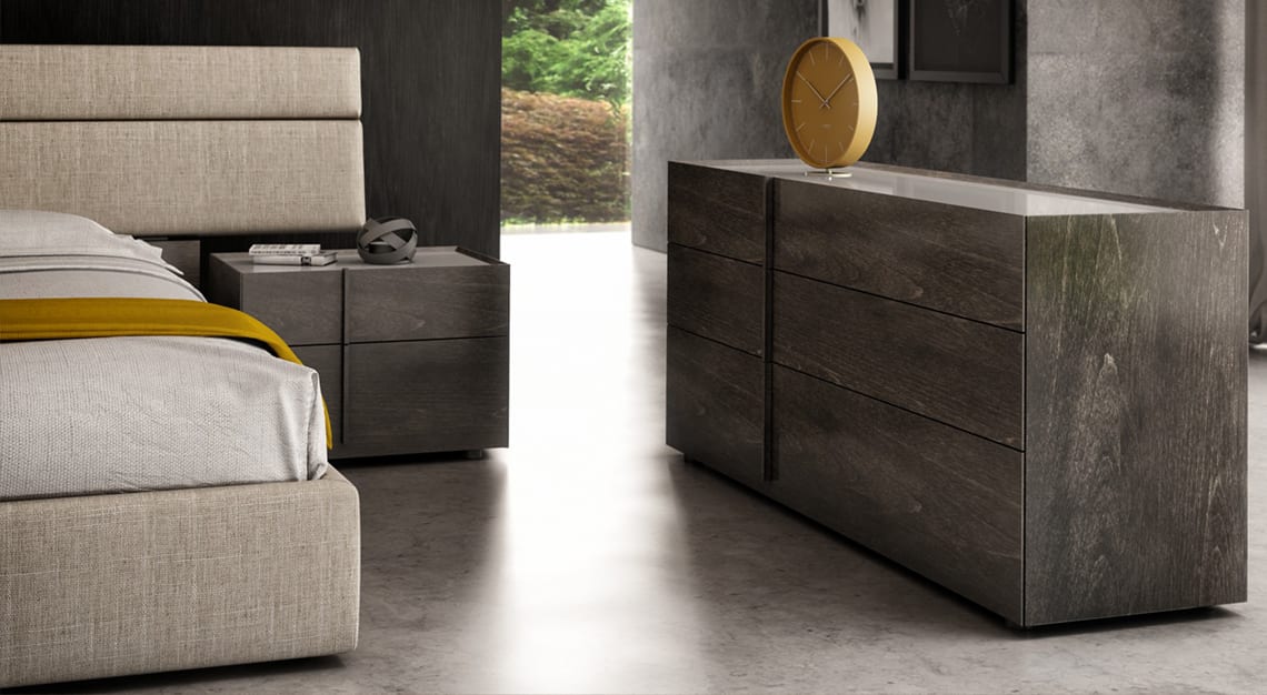 A modern bedroom featuring a beige upholstered bed, a dark wood nightstand, and a dark wood dresser with a mustard yellow clock. Green foliage is visible outside a large opening in the background.