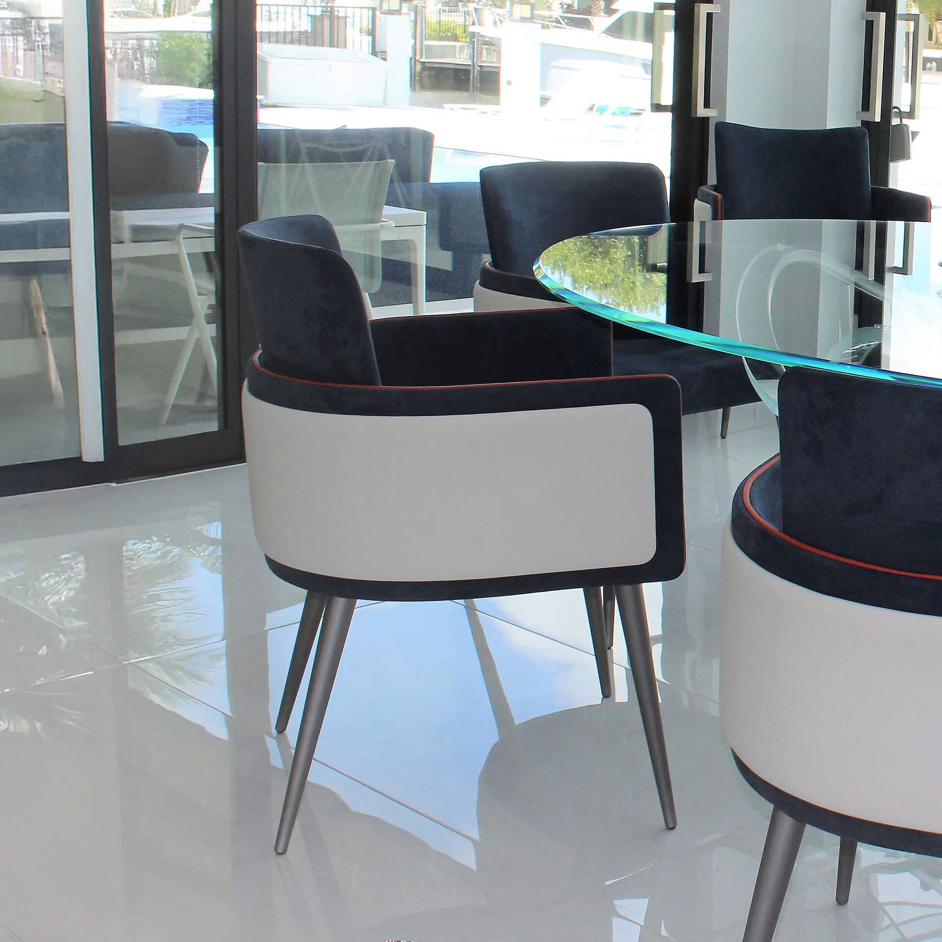Modern dining area with dark blue and cream chairs featuring orange piping, arranged around a glass table with a blue edge. Large windows overlook a bright blue pool and a waterfront with boats.