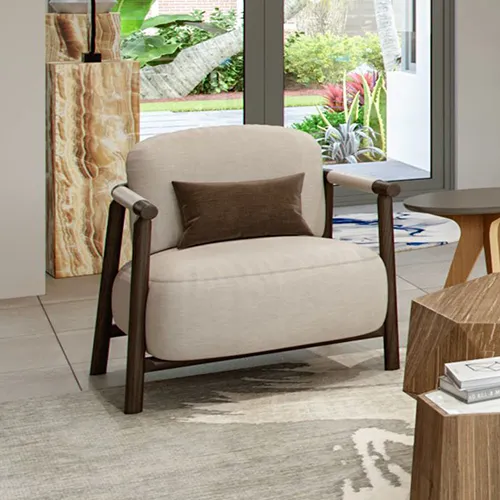A light beige armchair with a dark wooden frame and a brown pillow sits on a patterned rug in a modern living room, with a garden visible through a large window.