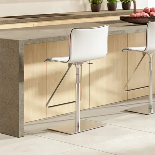 Two white bar stools with silver bases at a modern kitchen island with a gray countertop and wooden cabinetry. Potted plants and apples are on the counter.