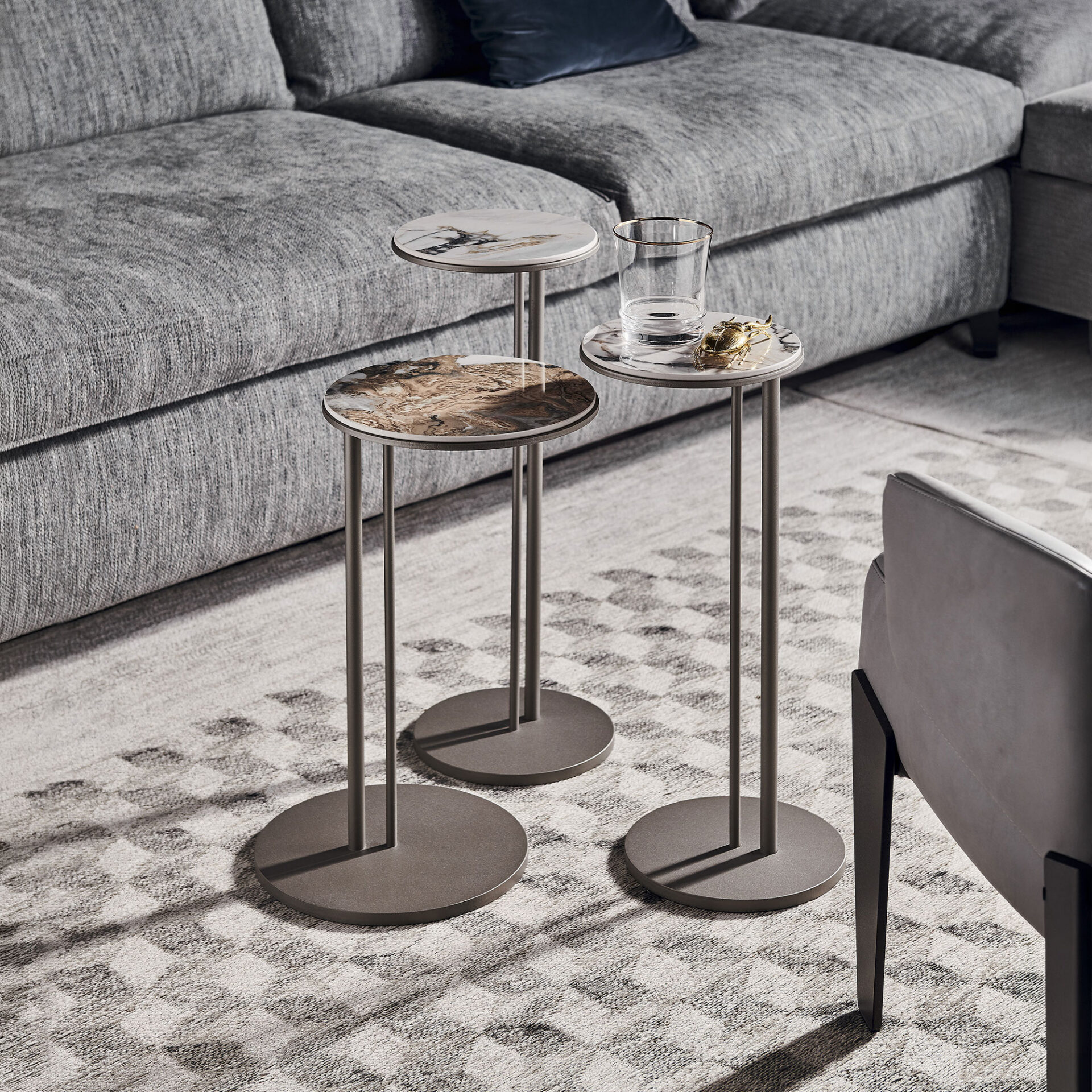 Three modern, round side tables with marble tops and dark metallic bases, next to a grey sectional sofa on a patterned rug. One table holds a gold-rimmed glass and a golden beetle figurine.