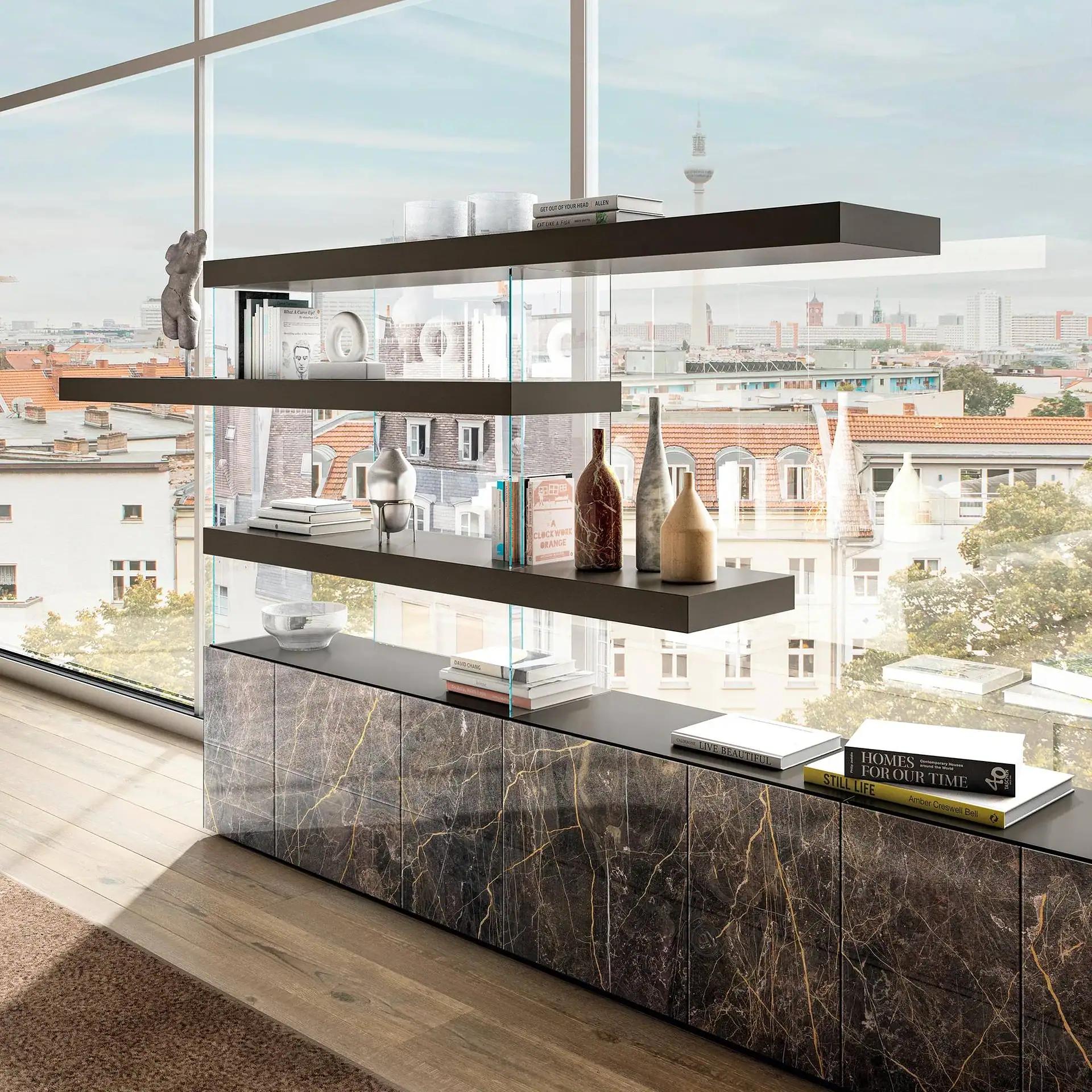 A modern living space with a large window overlooking a city. A dark marble-effect cabinet with gold veins supports floating shelves filled with books and decorative vases.