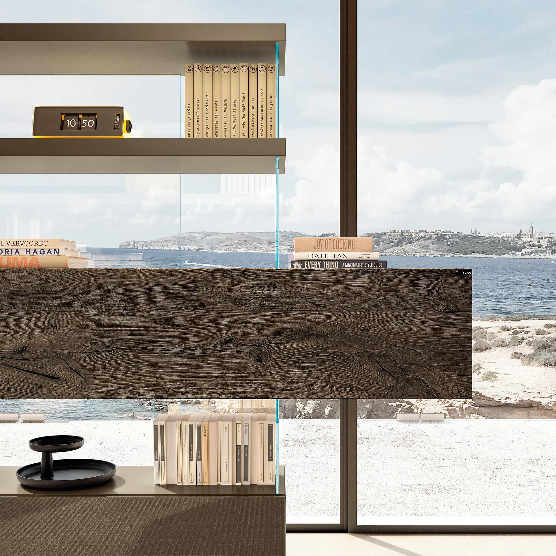 A modern shelving unit with books and a flip clock, set against a large window overlooking a blue sea, rocky coast, and distant town.